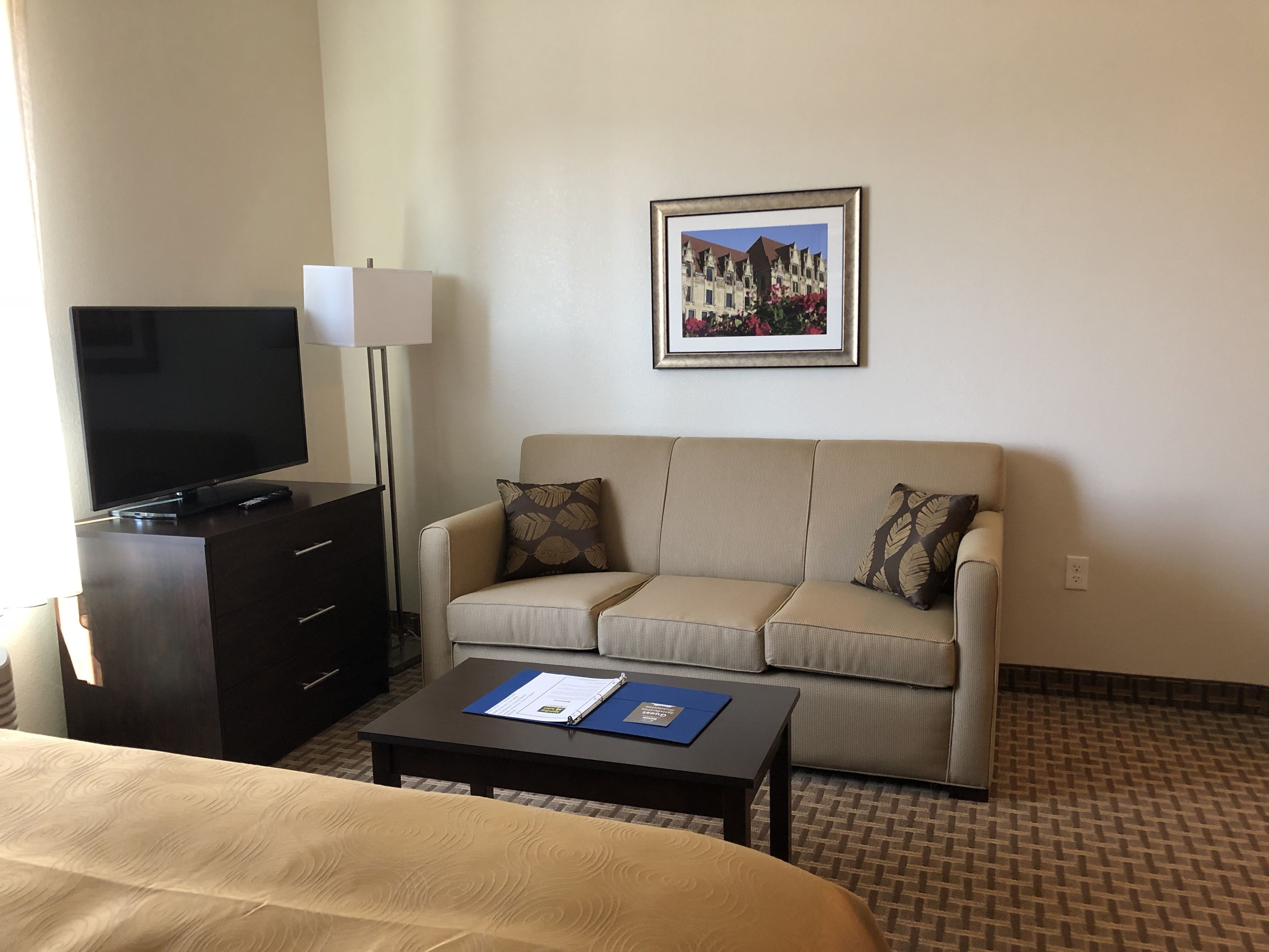 A hotel room with a bed, a TV on a dresser, a sofa with patterned cushions, a coffee table with a folder, and a framed picture on the wall.