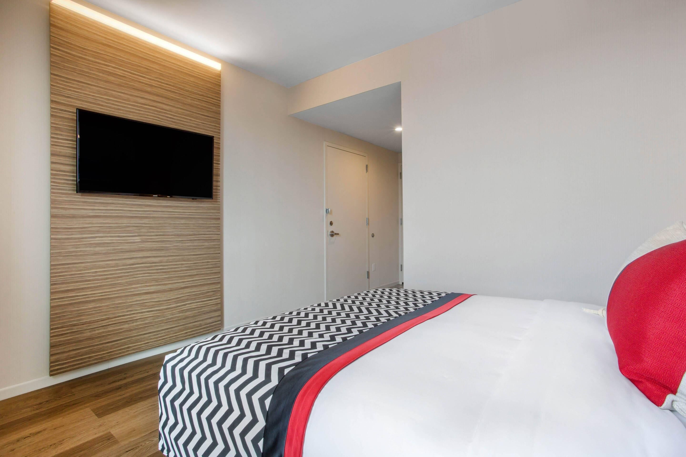 A modern bedroom with a flat-screen TV mounted on a wooden panel wall, a bed with a black and white zigzag patterned bedspread, and a white pillow.