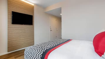 A modern bedroom with a flat-screen TV mounted on a wooden panel wall, a bed with a black and white zigzag patterned bedspread, and a white pillow.