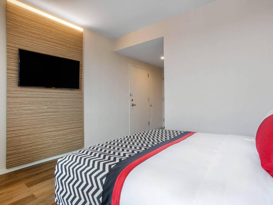 A modern bedroom with a flat-screen TV mounted on a wooden panel wall, a bed with a black and white zigzag patterned bedspread, and a white pillow.