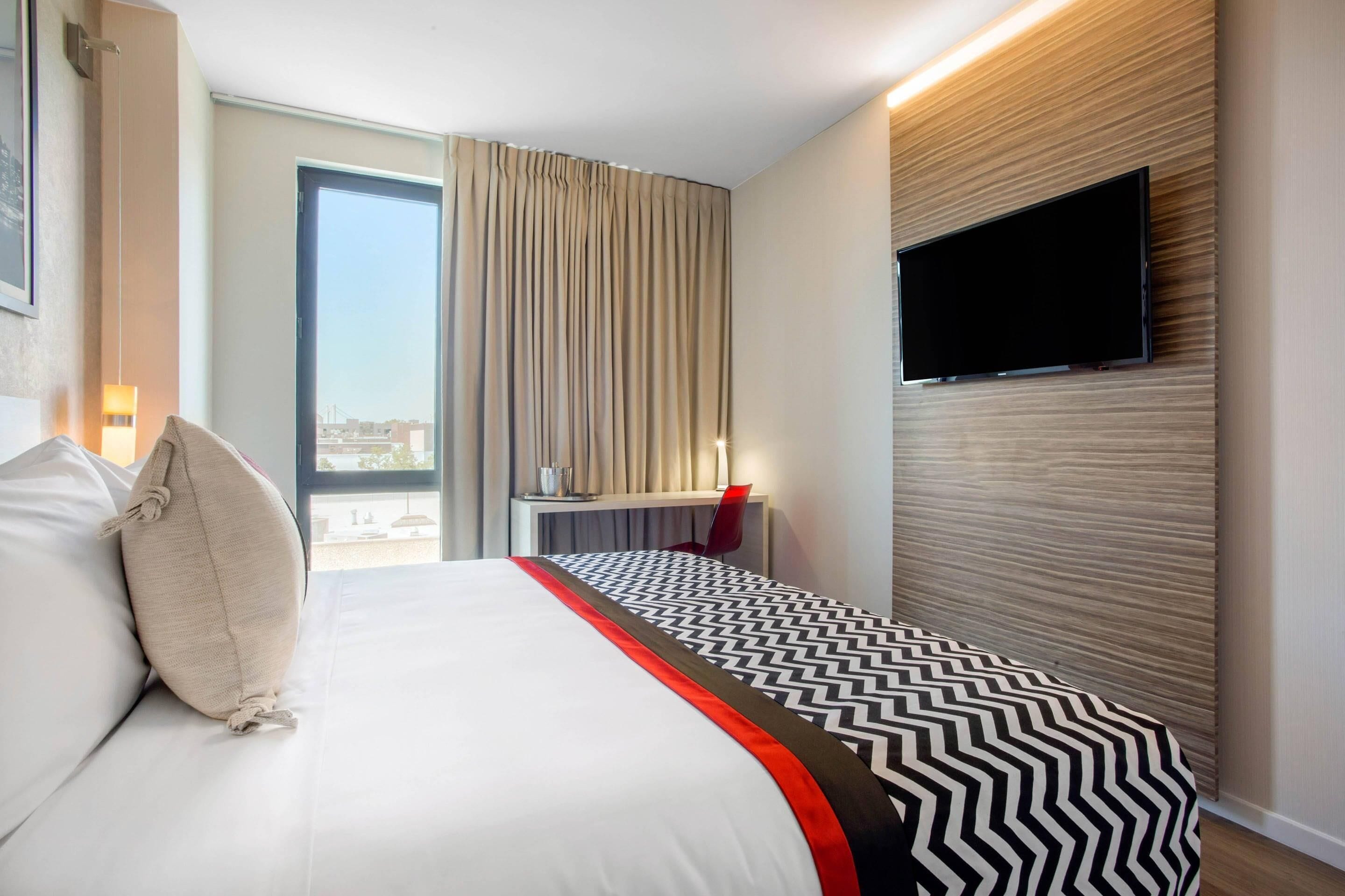 Habitación de hotel con una cama, televisión montada en la pared, escritorio con silla y ventana con cortinas.