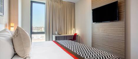 Habitación de hotel con una cama, televisión montada en la pared, escritorio con silla y ventana con cortinas.