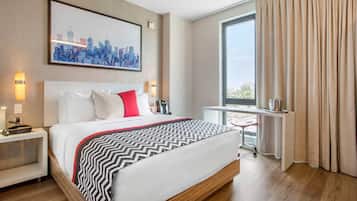 Quarto de hotel com uma cama grande, uma escrivaninha com cadeira, uma pintura de paisagem urbana e uma janela ampla com cortinas.