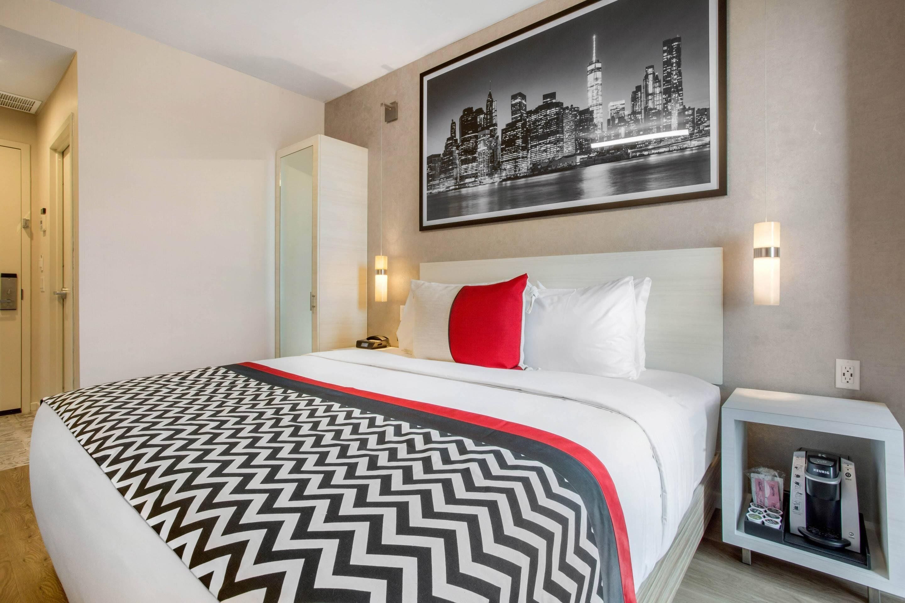 A hotel room with a bed, a nightstand with a coffee maker, and a cityscape picture on the wall.
