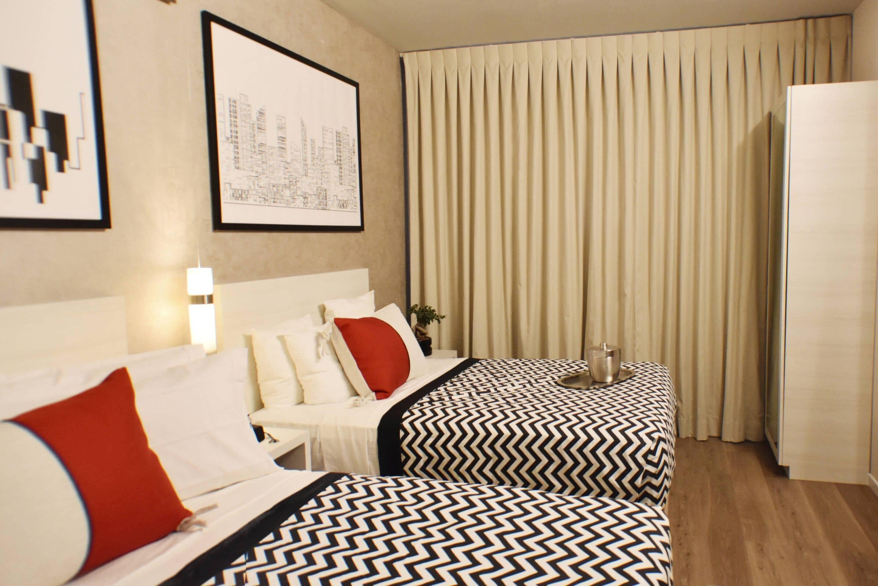 A hotel room with two beds, a large window with curtains, a wall-mounted artwork, and a bedside table with a lamp.