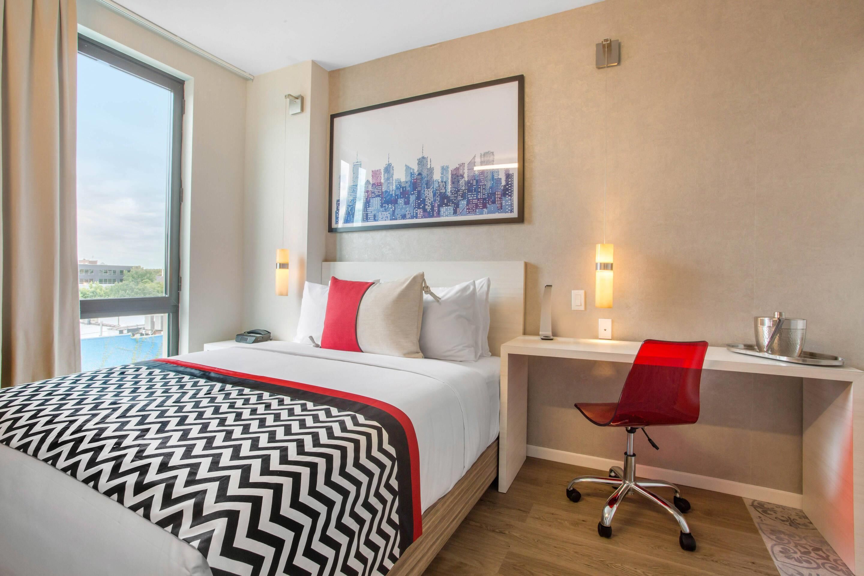 Ett hotellrum med en stor säng, ett skrivbord med en stol, ett konstverk med en stadsbild på väggen och ett fönster med utsikt.
