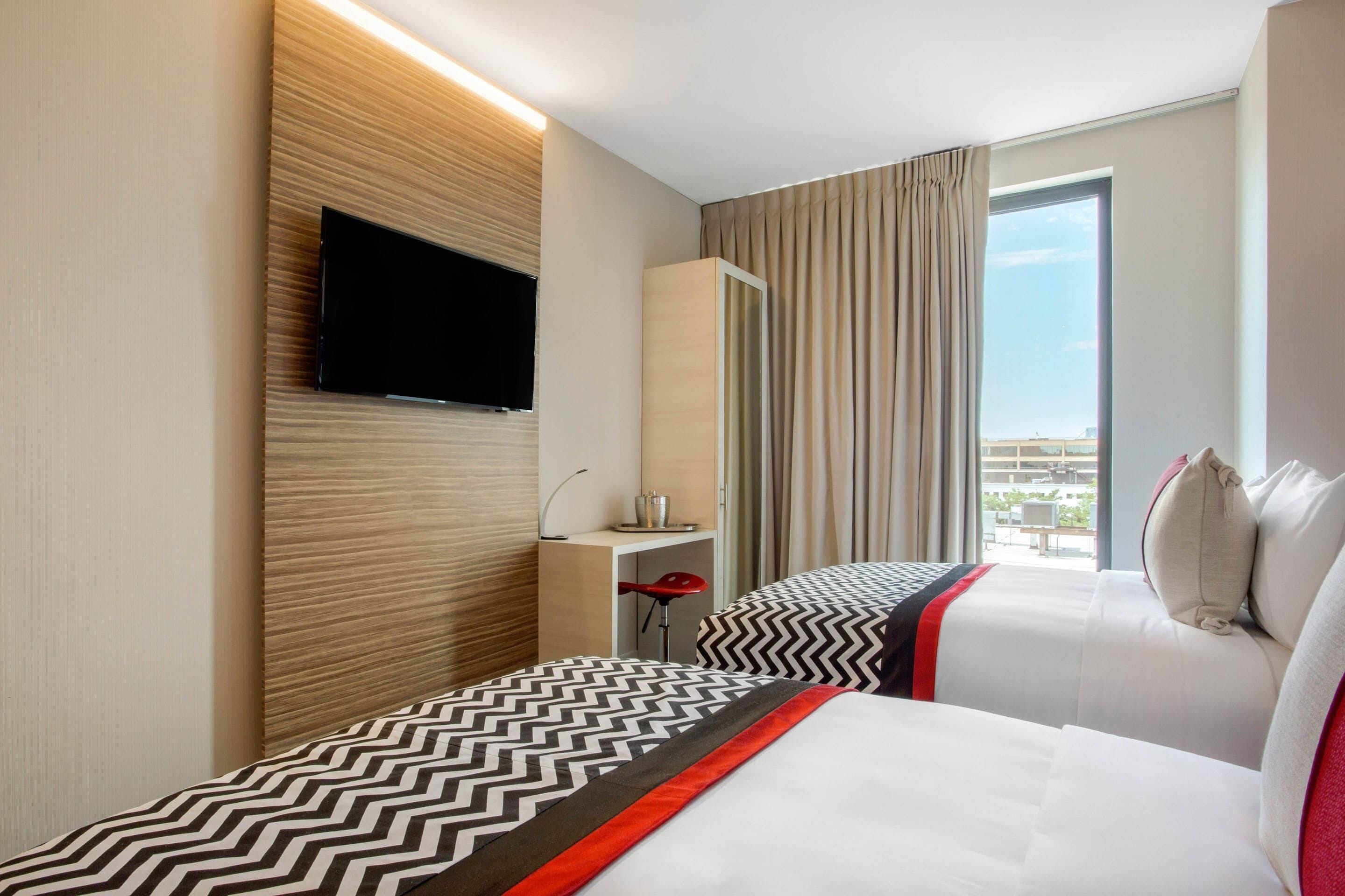 A hotel room with a bed, a TV mounted on the wall, a desk with a chair, and a window with curtains.