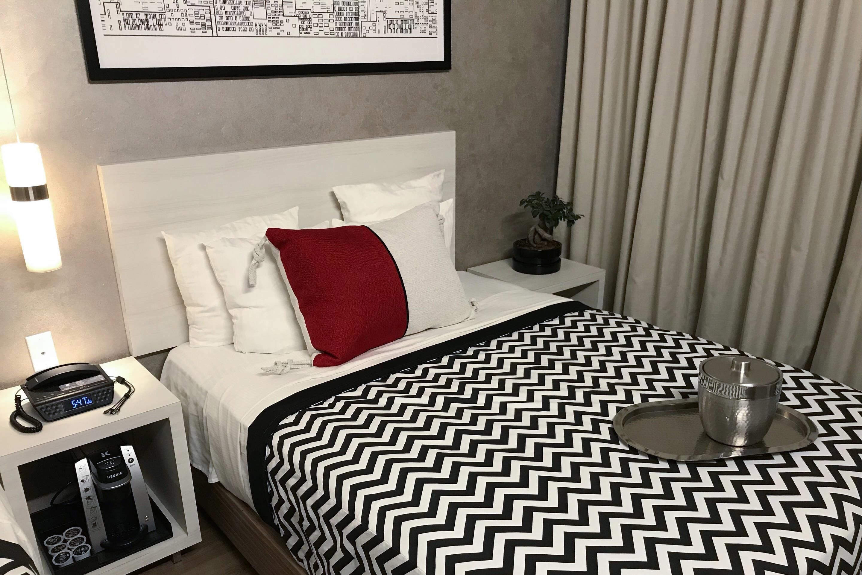 A neatly arranged hotel room with a bed, a bedside table with a phone and a coffee maker, a small plant, and a wall-mounted map.