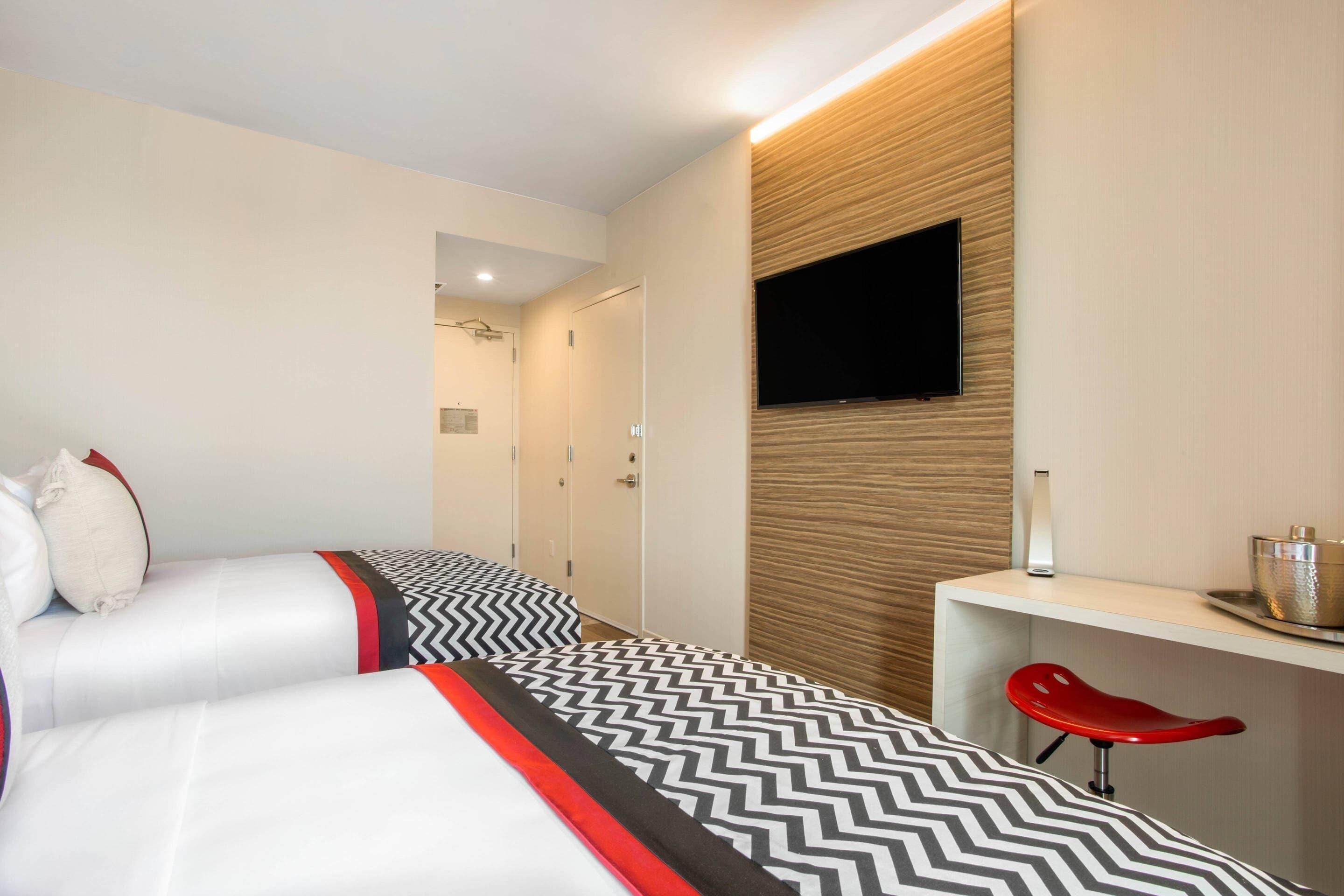 A hotel room with two beds, a TV mounted on the wall, a desk with a chair, and a small table with a bowl.