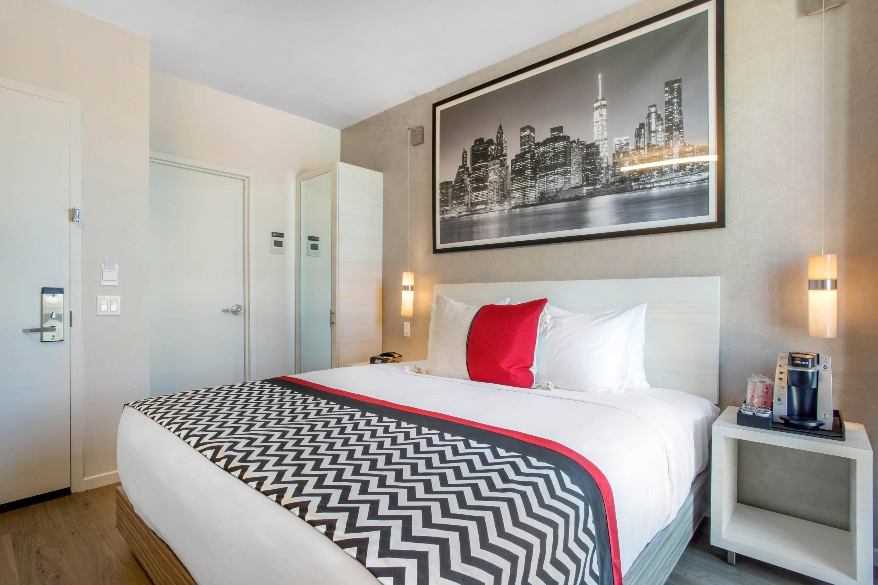 Una habitación de hotel con cama, un cuadro de paisaje urbano, una mesita de noche con cafetera y una puerta.