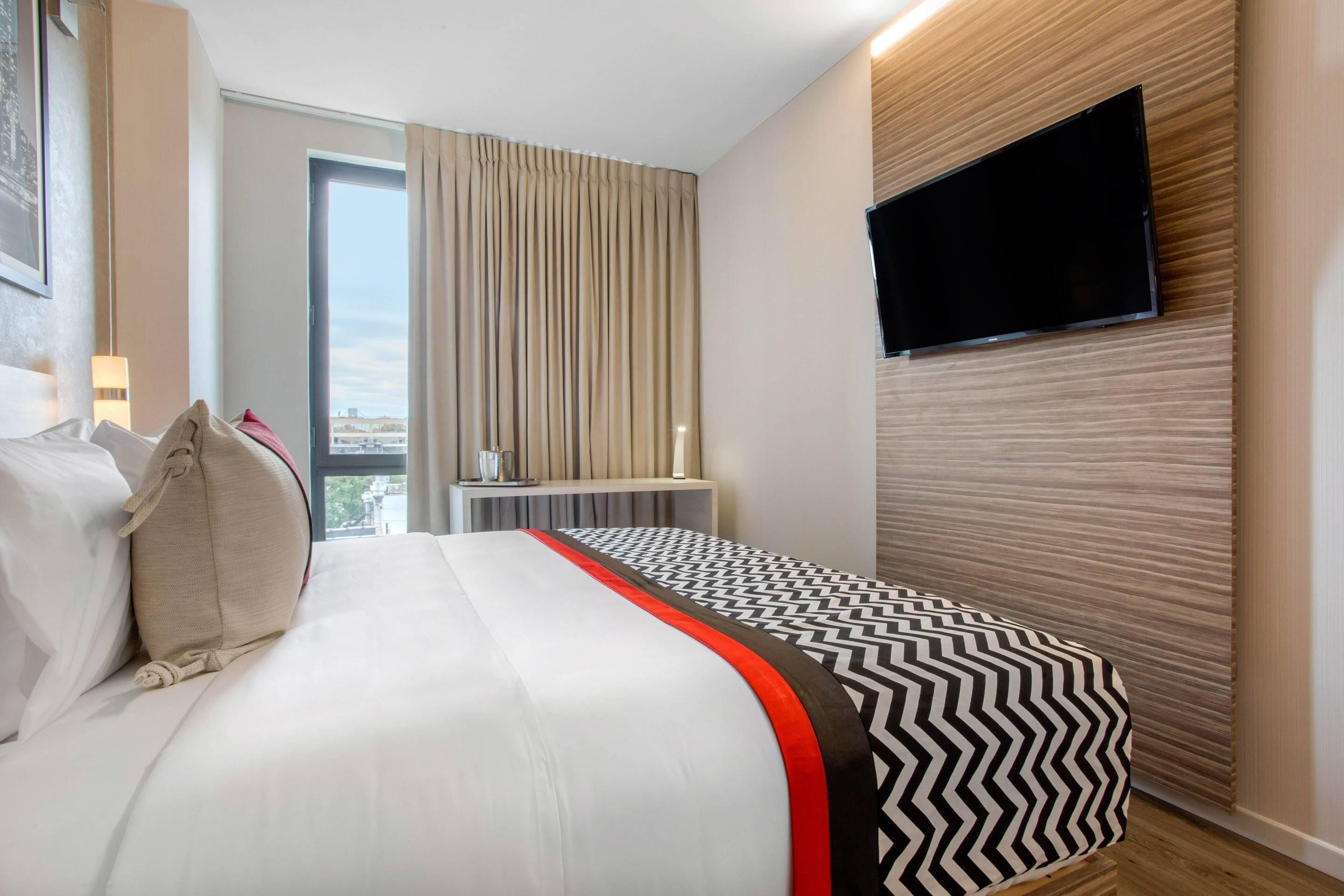 A hotel room with a large bed, a flat-screen TV mounted on the wall, a window with curtains, and a view of the city.