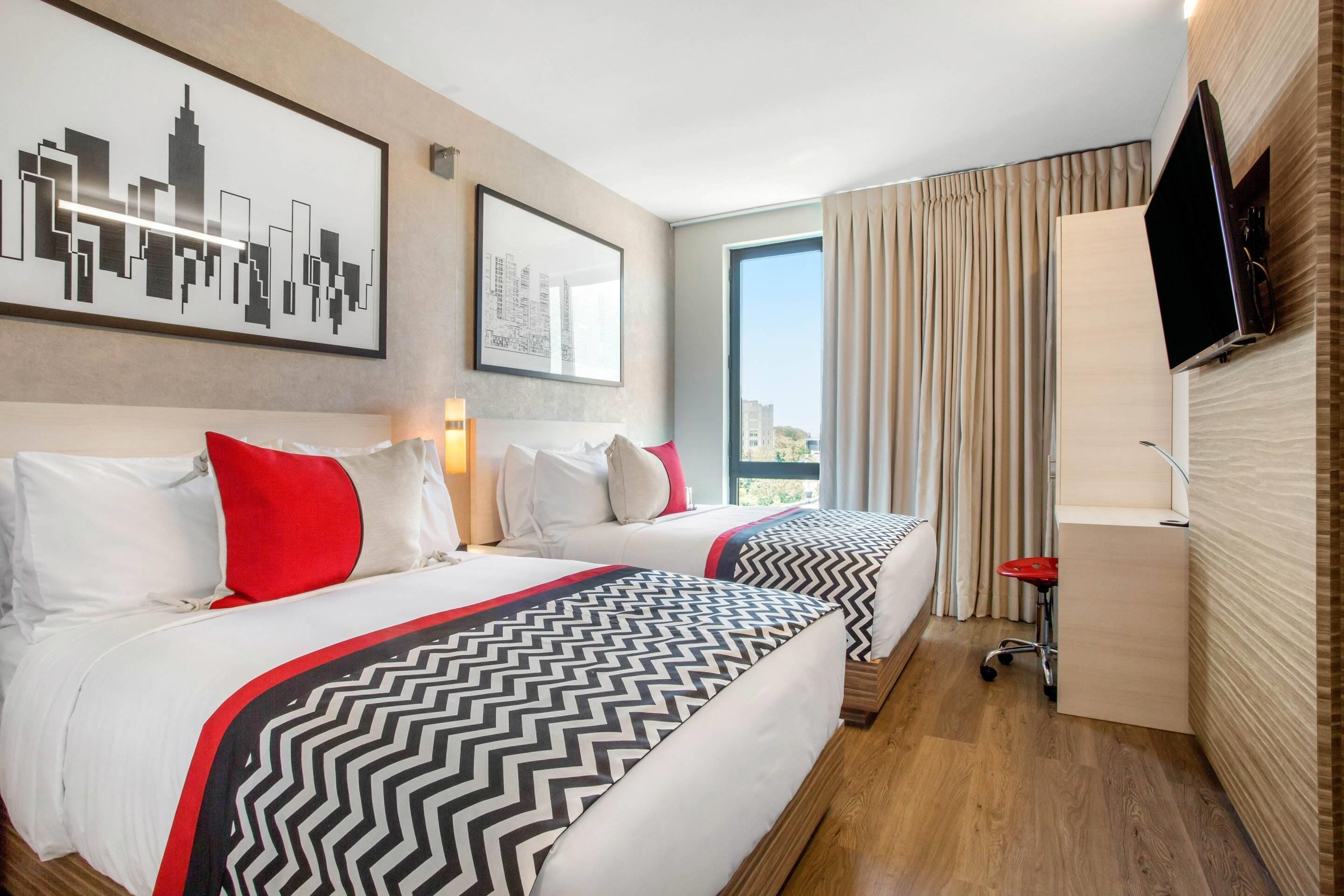 A hotel room with two beds, a TV, a desk, and a large window with curtains.