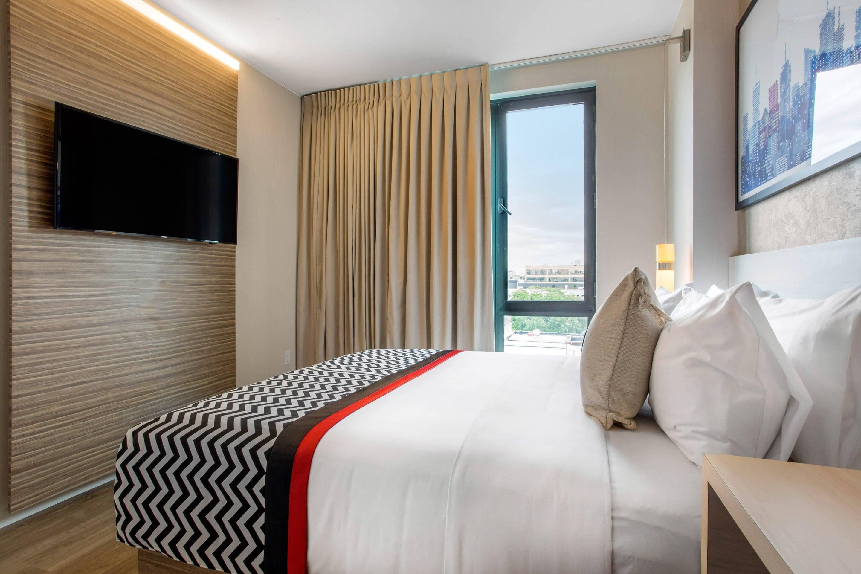 Een hotelkamer met een bed, een flatscreen televisie aan de muur, een raam met gordijnen en uitzicht op de stad.
