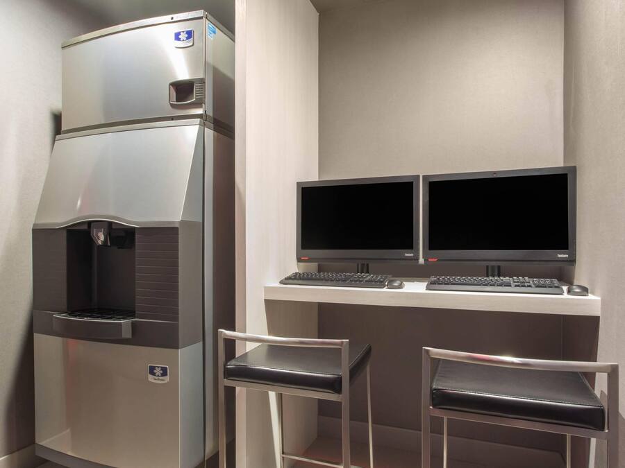 A compact kitchenette with a refrigerator, two monitors, a keyboard, and two chairs.
