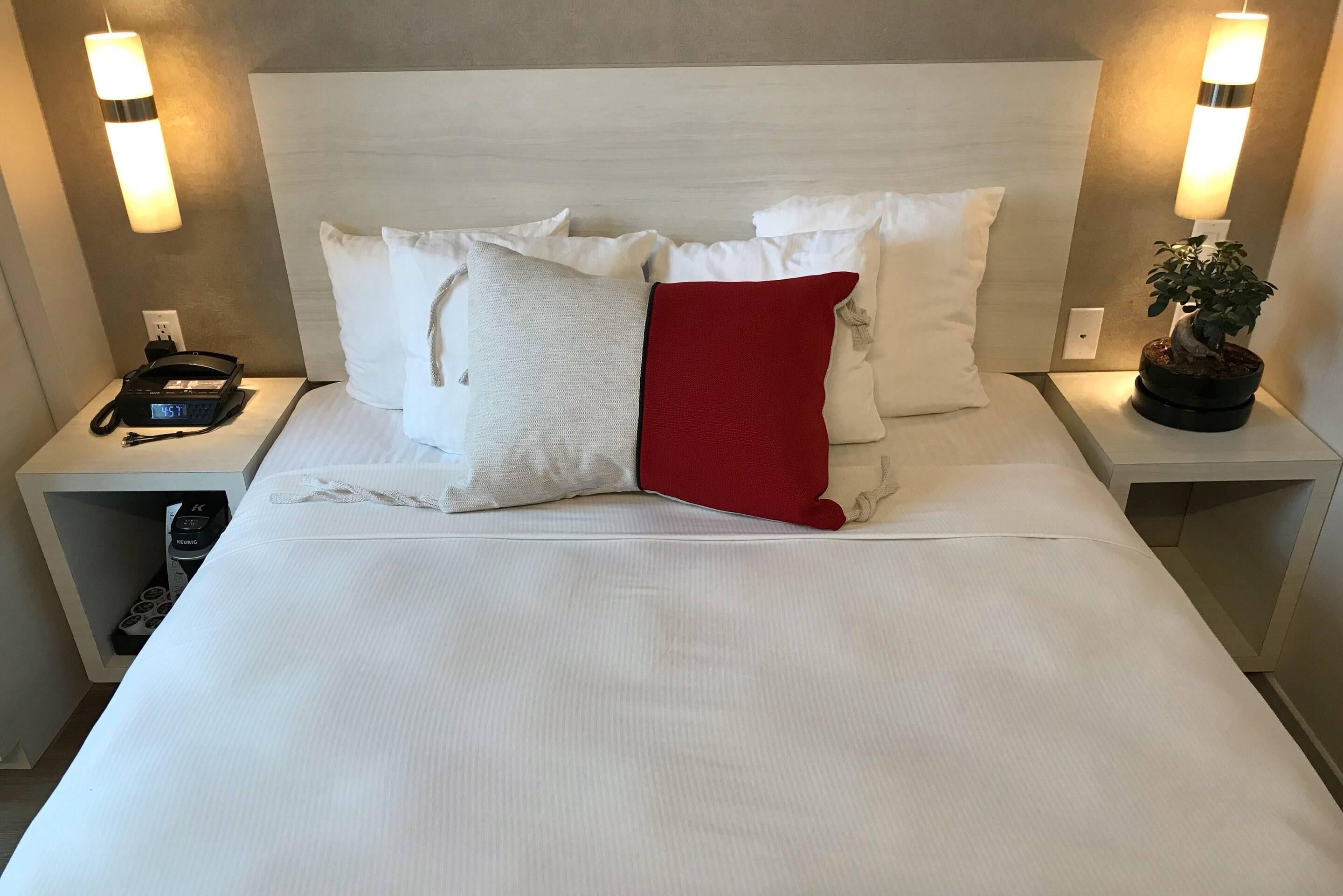 Een netjes opgemaakt bed met witte en rode kussens, een bedtelefoon en een kleine plant in een potje op het nachtkastje.