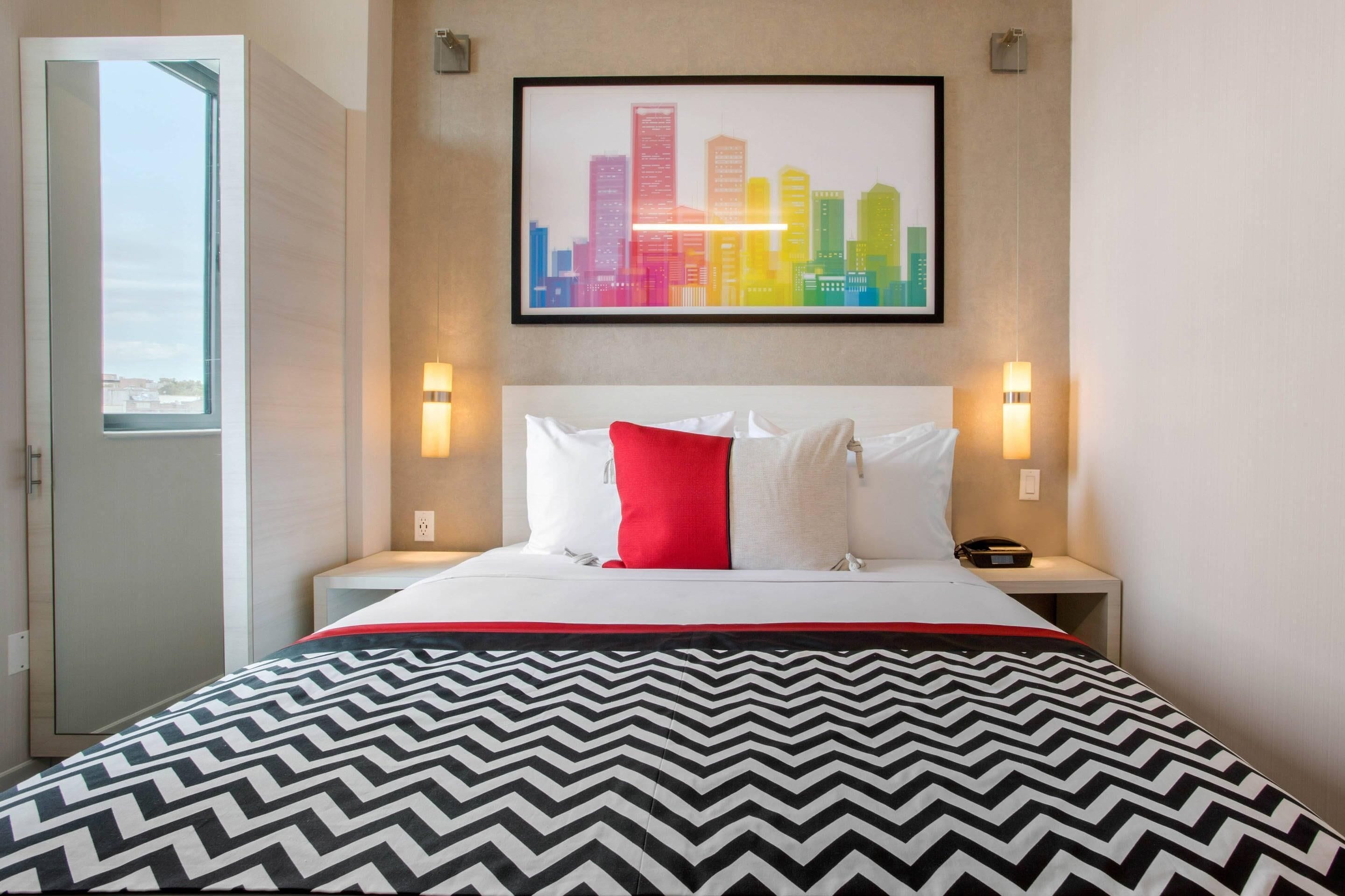 Une chambre d’hôtel avec un lit, une œuvre d’art représentant un paysage urbain accrochée au mur, des lampes de chevet et une fenêtre.