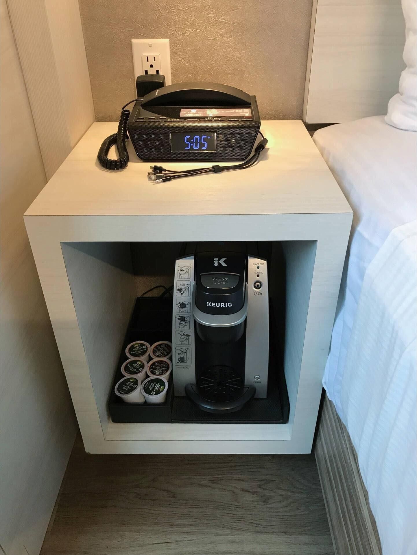 A hotel room with a bedside table that includes a digital clock and a coffee maker.