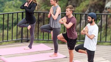 Tres personas practicando yoga sobre esterillas rosas en una terraza de madera con barandilla, rodeadas de follaje.