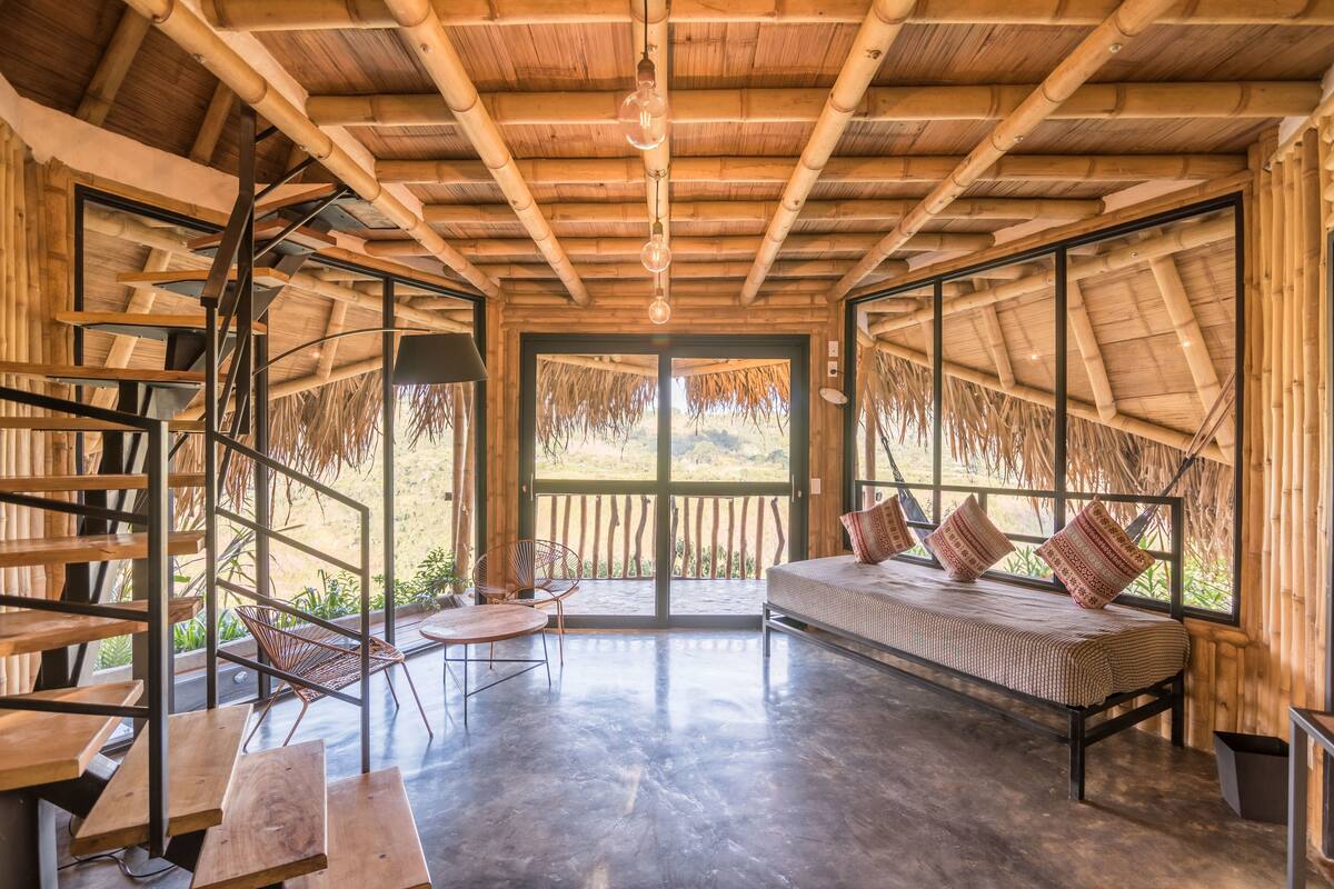 A spacious room with a large bed, a dining table, and chairs. There is a large window with a view of the outdoors.