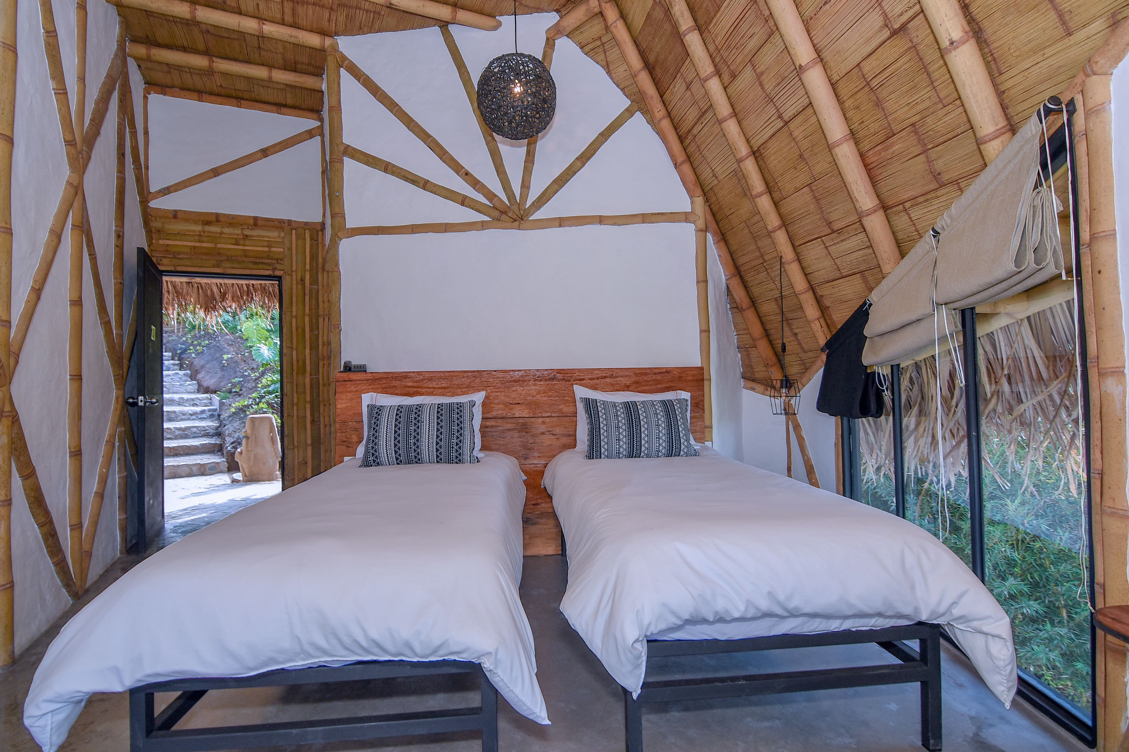 A room with two beds, a wooden ceiling, and a view of greenery outside.