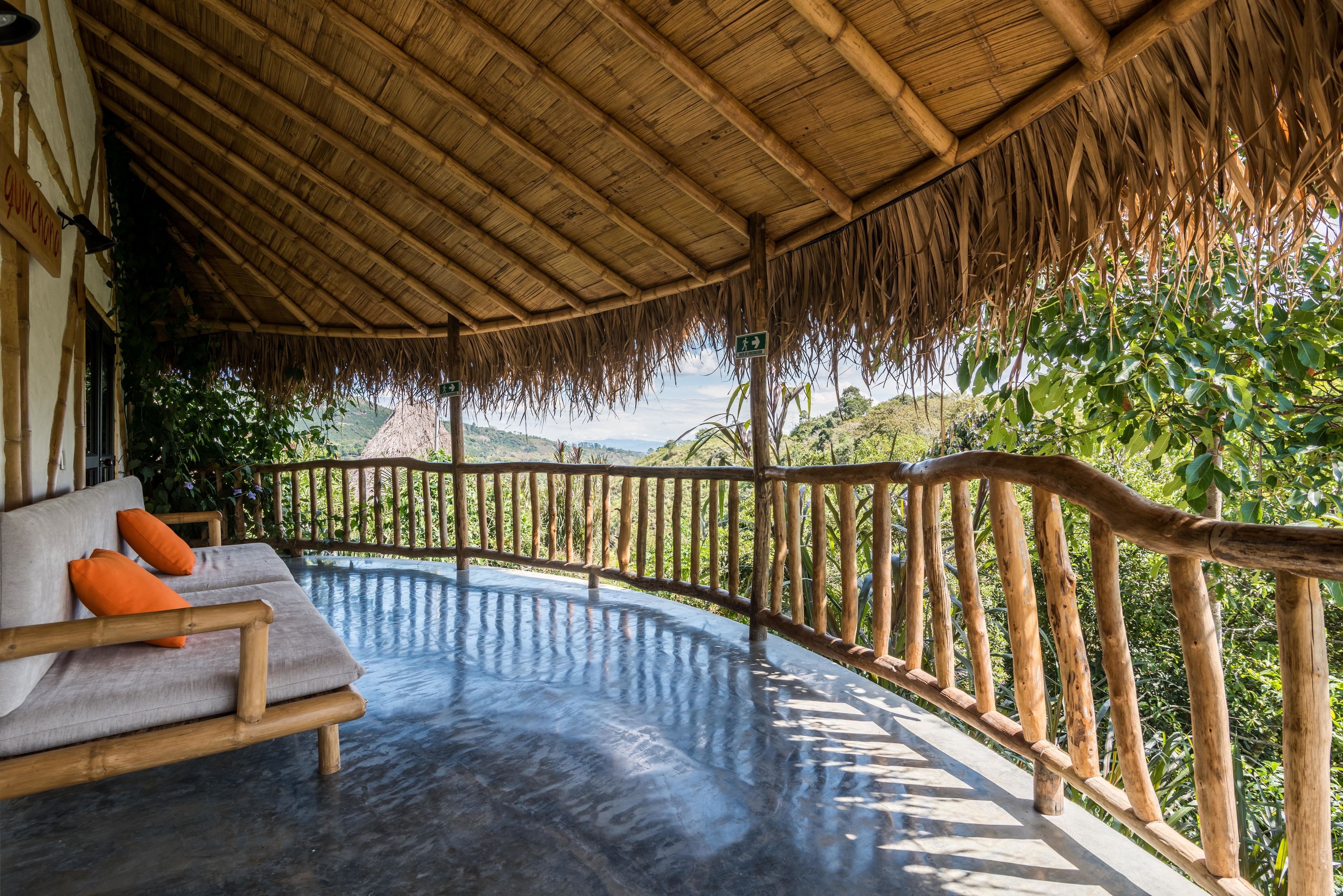 A covered outdoor seating area with a wooden bench and cushions, a thatched roof, and a view of lush greenery and hills.