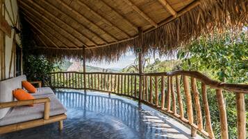 A covered outdoor seating area with a wooden bench and cushions, a thatched roof, and a view of lush greenery and hills.