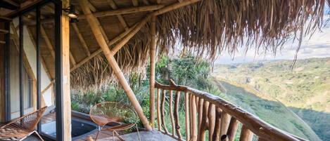 Un balcón de madera con techo de paja, suelo de piedra y vista a un valle verde.