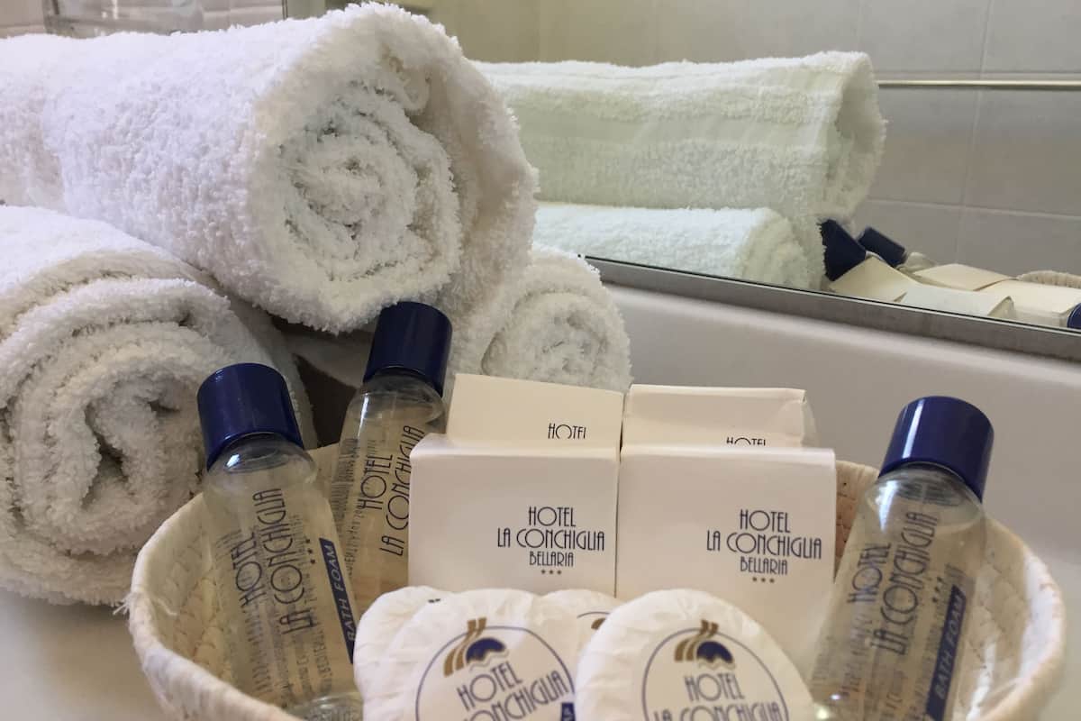 Une salle de bain avec des serviettes blanches roulées, des flacons de mousse de bain et des produits de toilette à l’effigie de l’hôtel.
