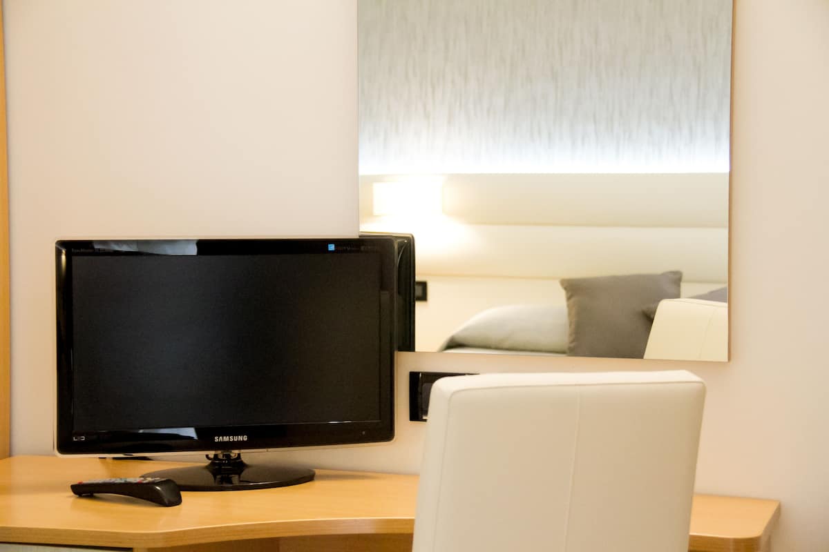 Une chambre d’hôtel avec un écran Samsung posé sur un bureau, une télécommande, une chaise blanche et un grand miroir qui reflète un lit avec des oreillers.