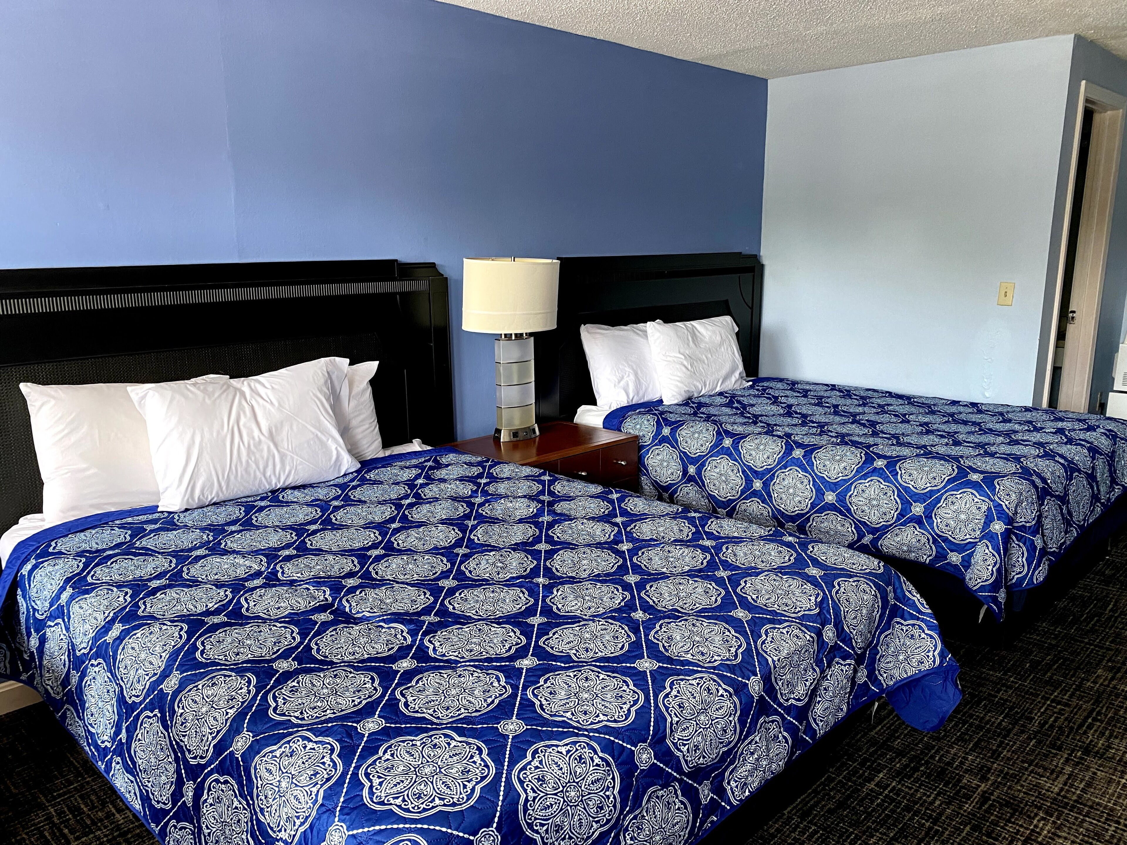 A hotel room with two beds, each with a blue patterned bedspread and white pillows. There is a nightstand with a lamp between the beds.