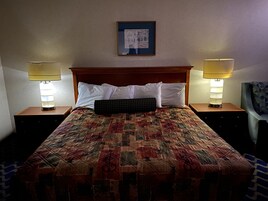 Una cama bien hecha con colcha, dos lámparas de noche, un cuadro enmarcado en la pared y una silla con estampado geométrico.