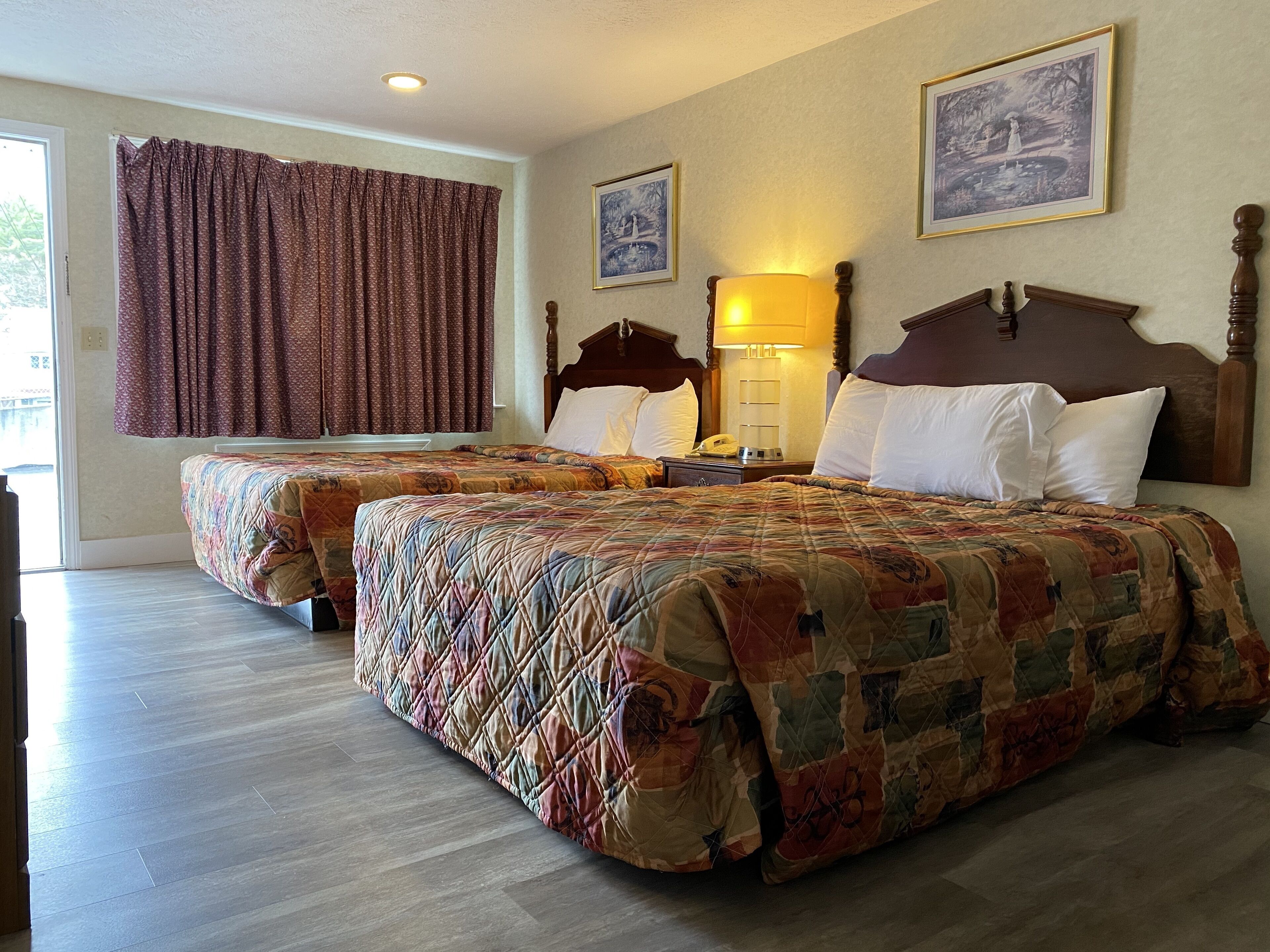 Habitación de hotel con dos camas tamaño queen, un ventanal grande con cortinas, cabecera de madera, lámpara de noche y cuadros en la pared.