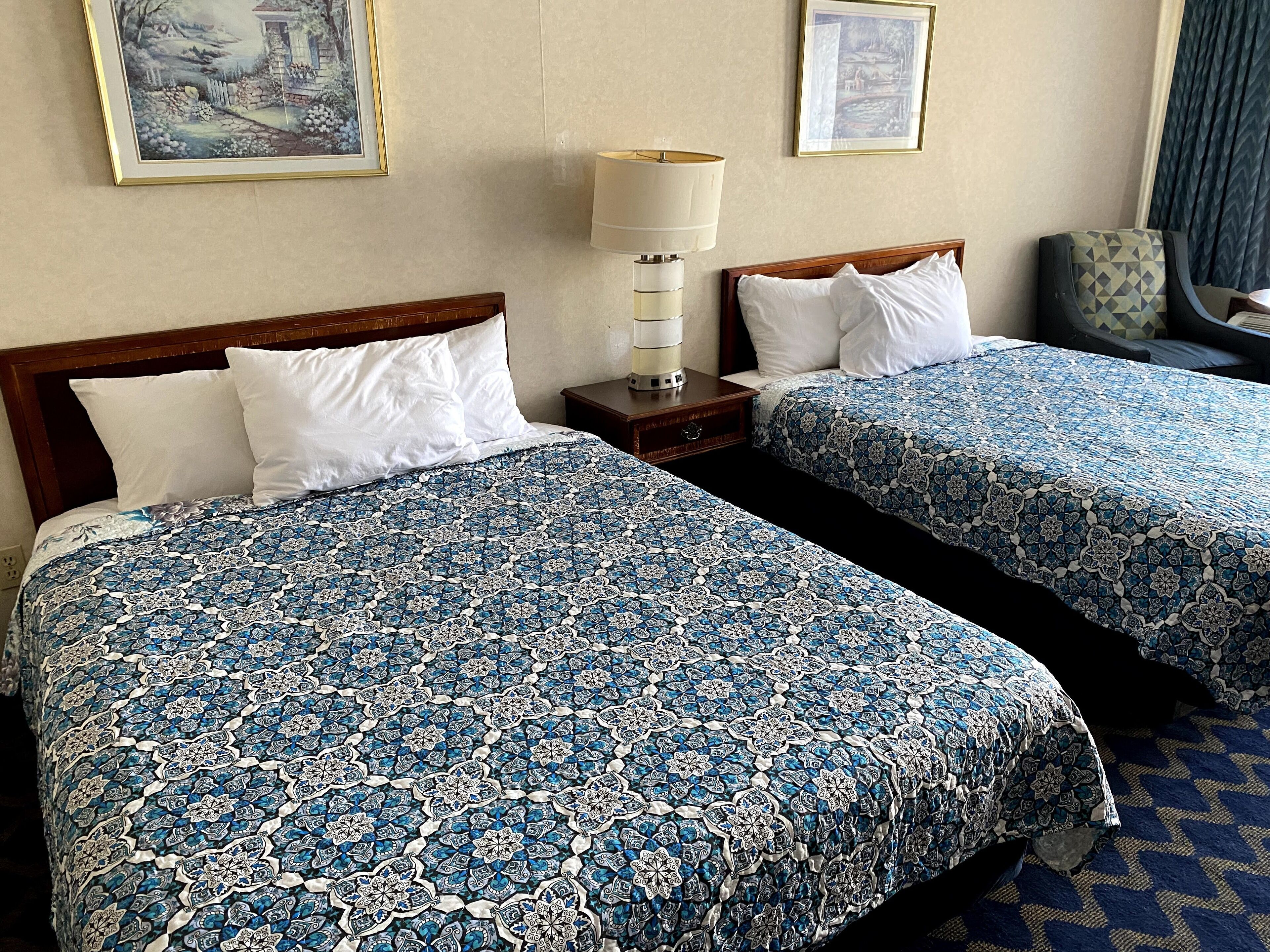 Une chambre d’hôtel avec deux lits, chacun recouvert d’une couvre-lit à motifs, une table de chevet avec une lampe et des œuvres d’art encadrées accrochées aux murs.