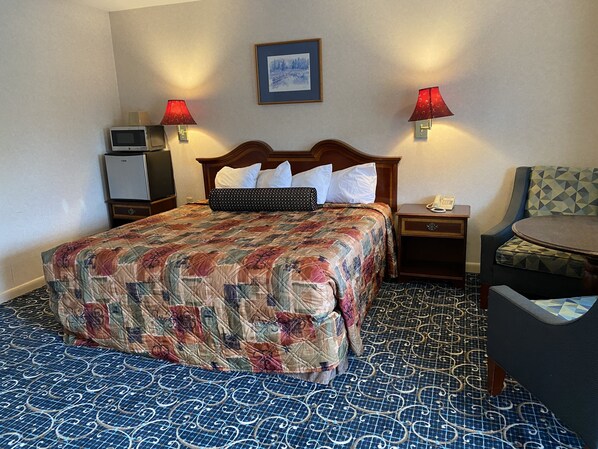 A hotel room with a large bed, two bedside lamps, a small table, a chair, and a microwave.