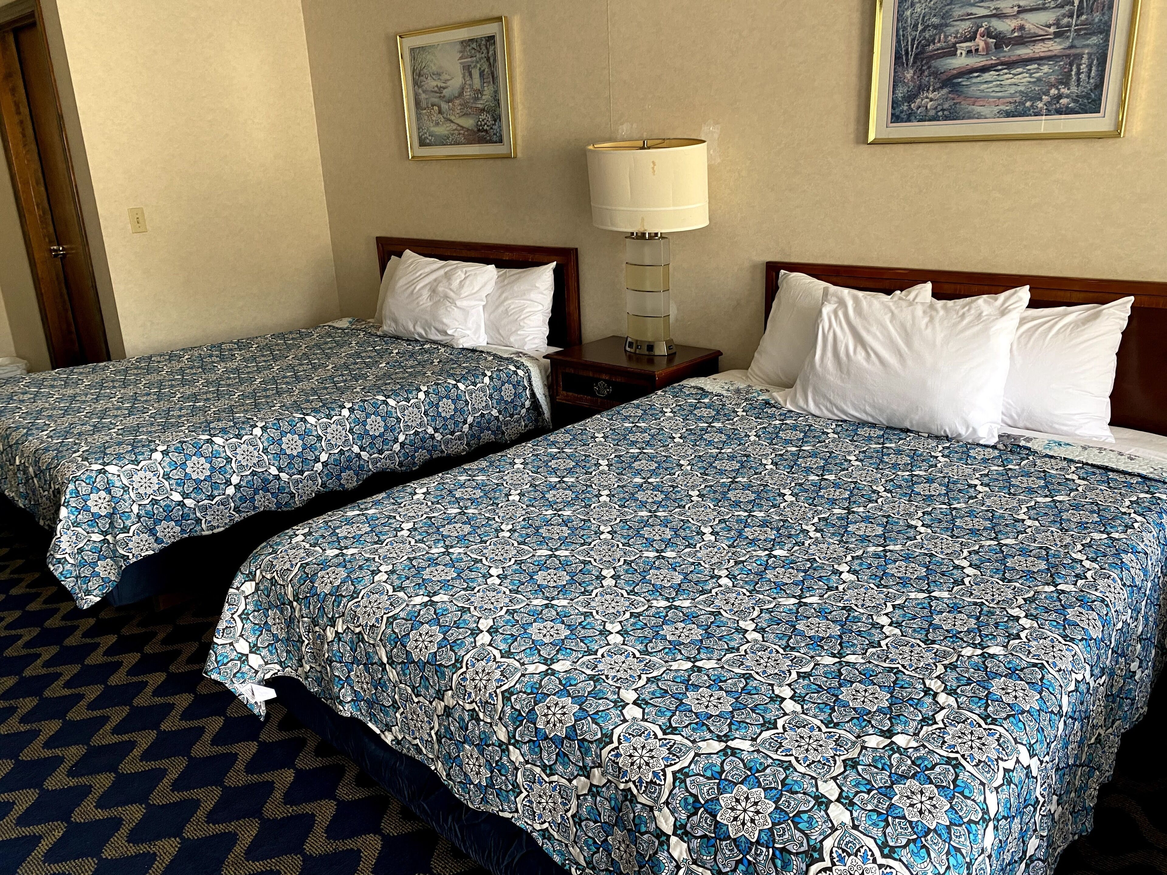 Two double beds with matching blue patterned bedspreads, a bedside table with a lamp, and a framed picture on the wall.