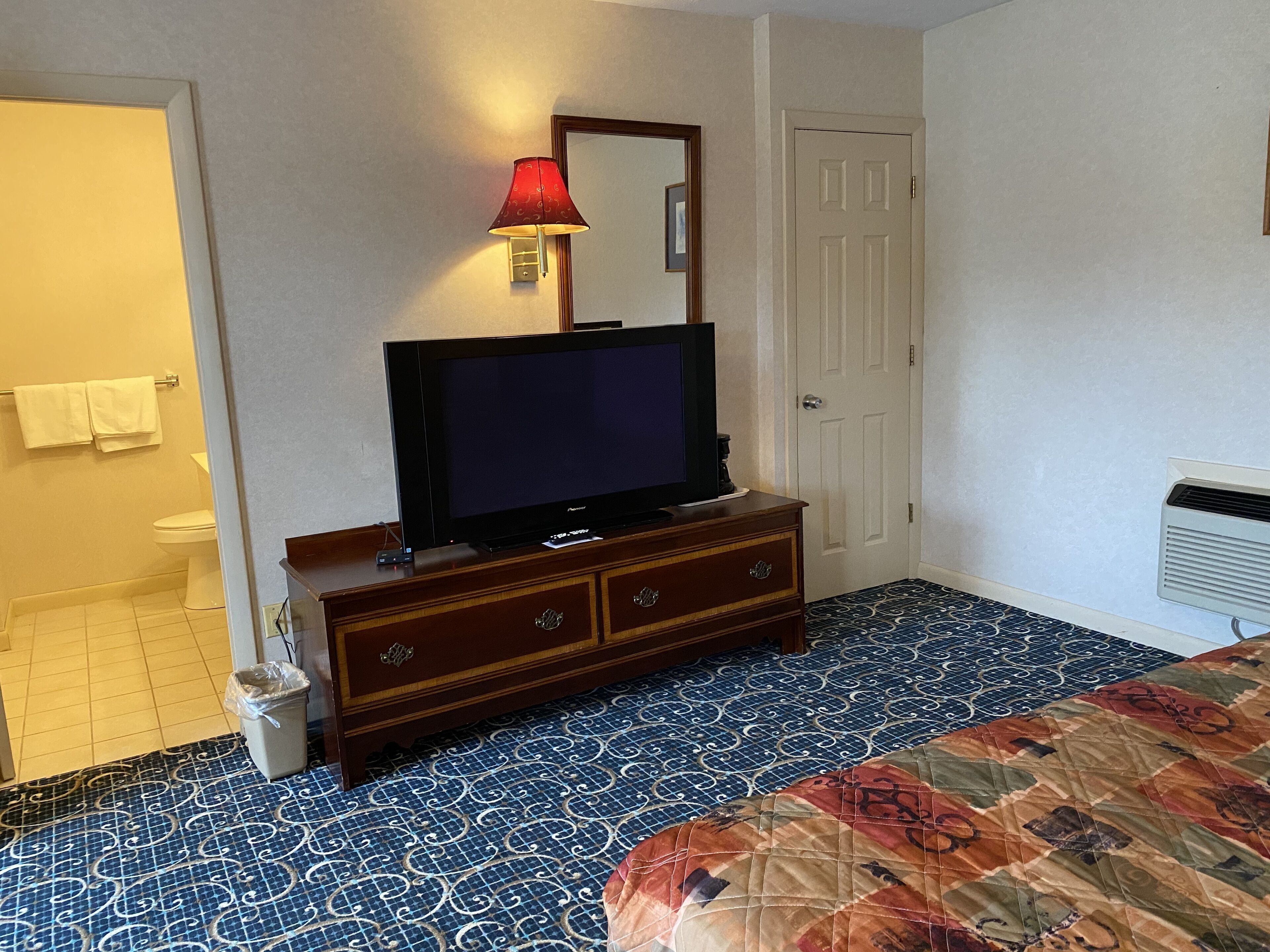 A hotel room with a television on a wooden dresser, a red lamp, a patterned floor, and a door.