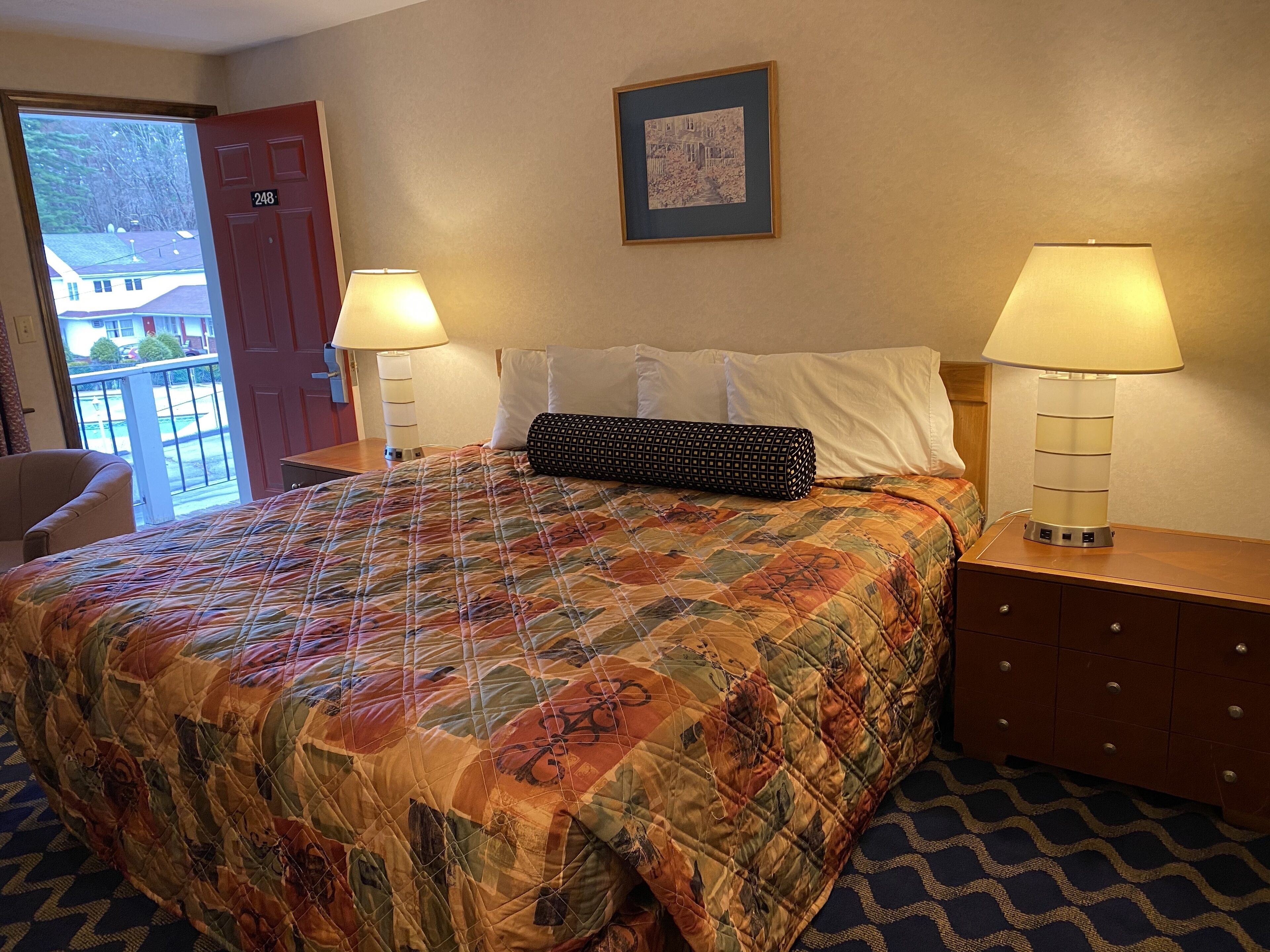 A hotel room with a large bed, two bedside lamps, a door, a framed picture on the wall, and a view of the outside.