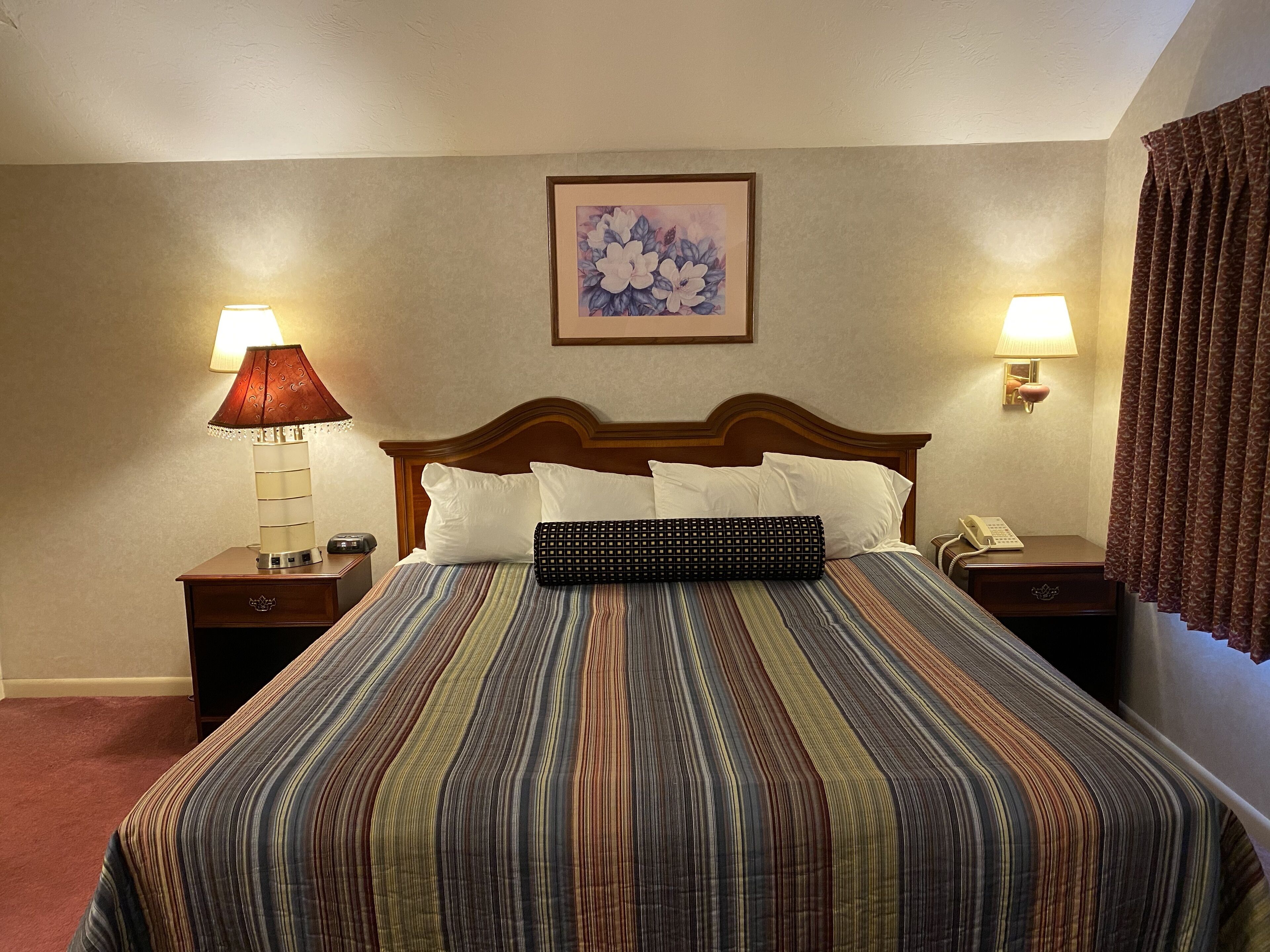 Une chambre d’hôtel avec un grand lit, deux lampes de chevet, un téléphone et une œuvre d’art florale encadrée accrochée au mur.
