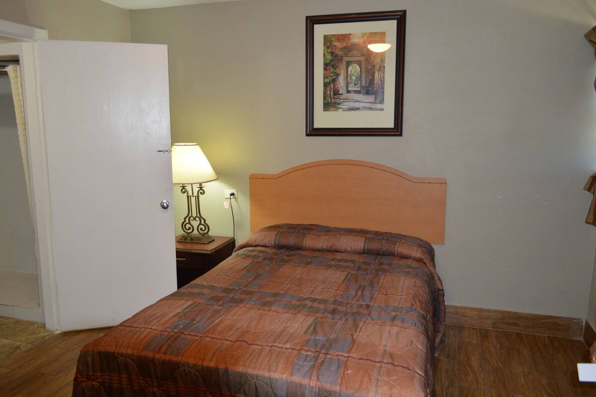 Une chambre à coucher avec un lit, une table de chevet avec une lampe, une porte et un tableau encadré au mur.