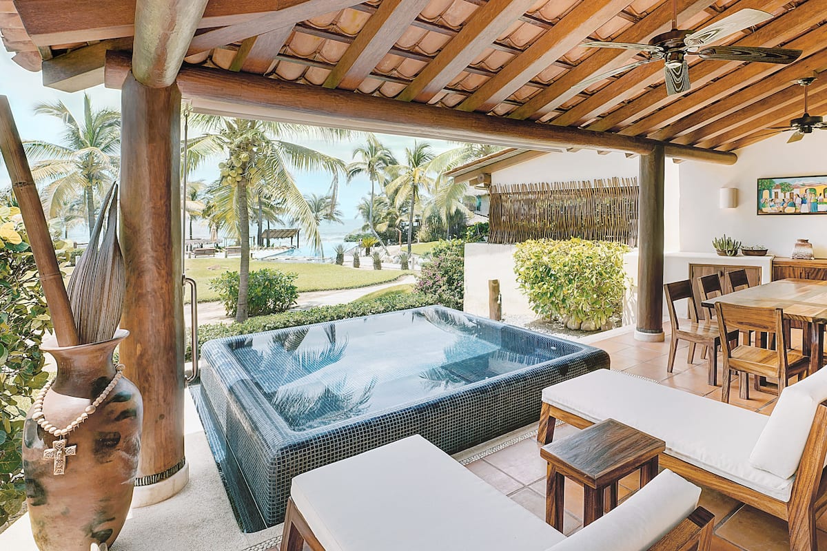 Un área de piscina con jacuzzi, mobiliario de madera y vistas a palmeras y al océano.