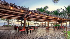 Una terraza de madera con mesas y sillas, palmeras y una playa al fondo.