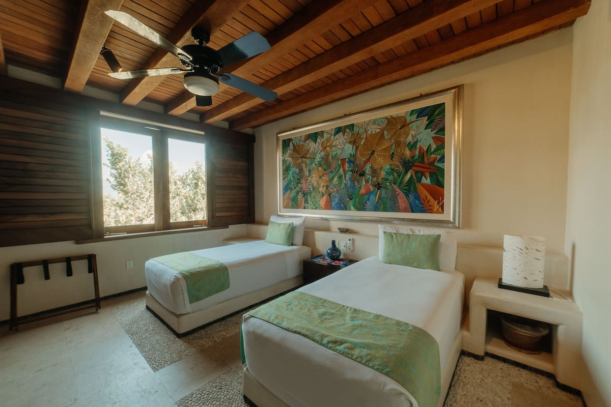 Habitación de hotel con dos camas, ventilador de techo, un cuadro abstracto grande y una ventana con vistas a árboles.
