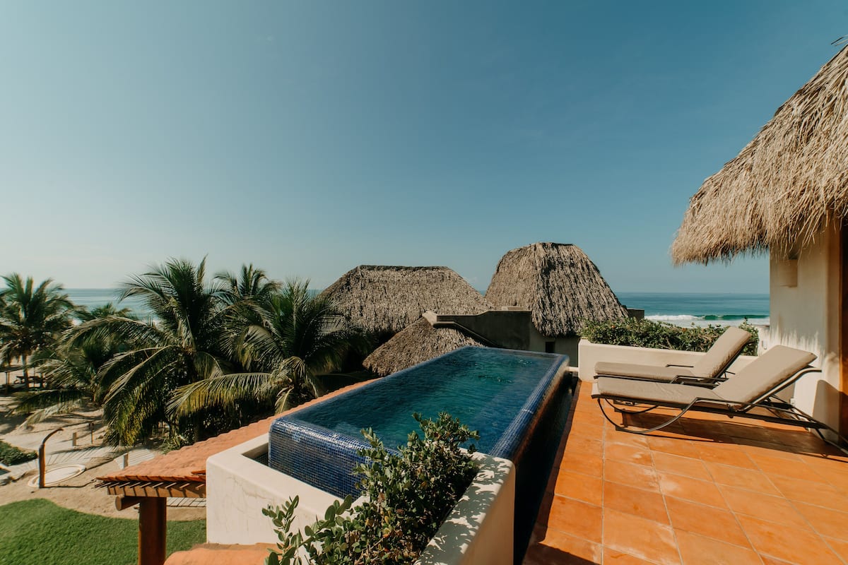 Piscina infinita en la azotea con vistas al océano, sillas de descanso y estructuras de techo de paja.