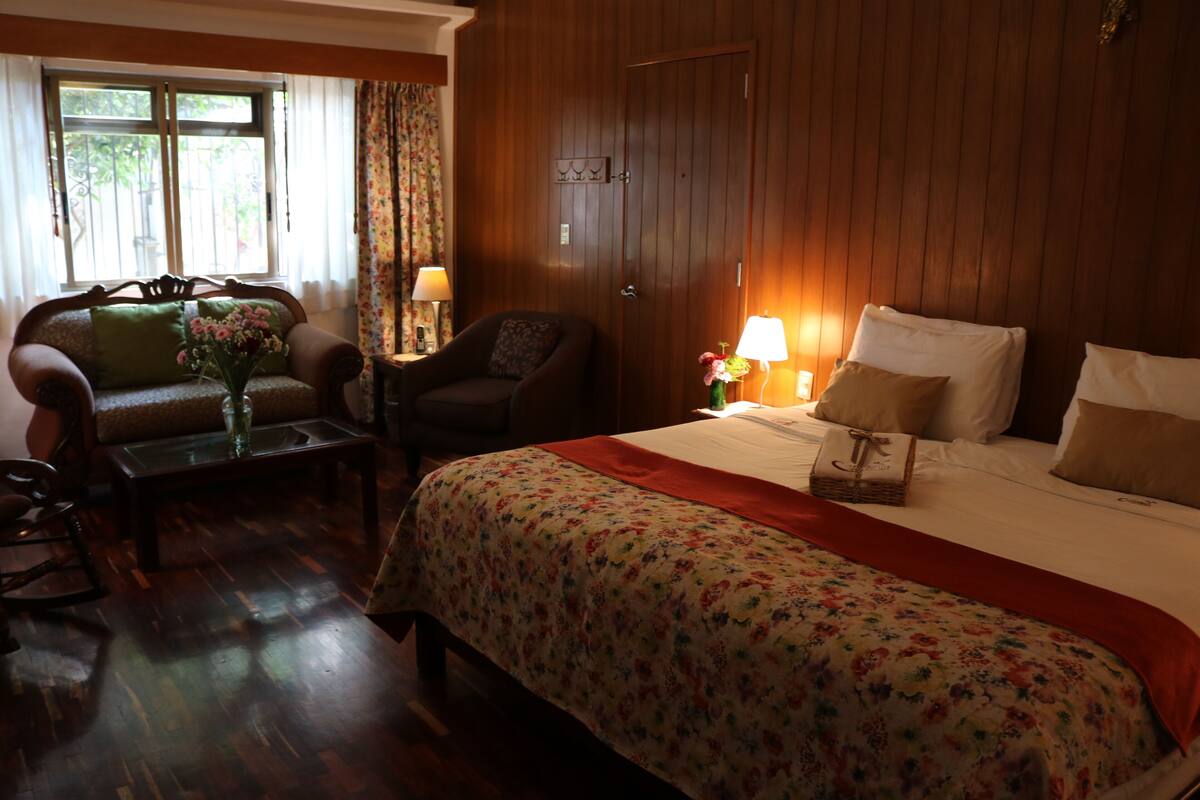 A hotel room with a wooden floor, a floral bedspread, a wooden door, and a window with curtains.