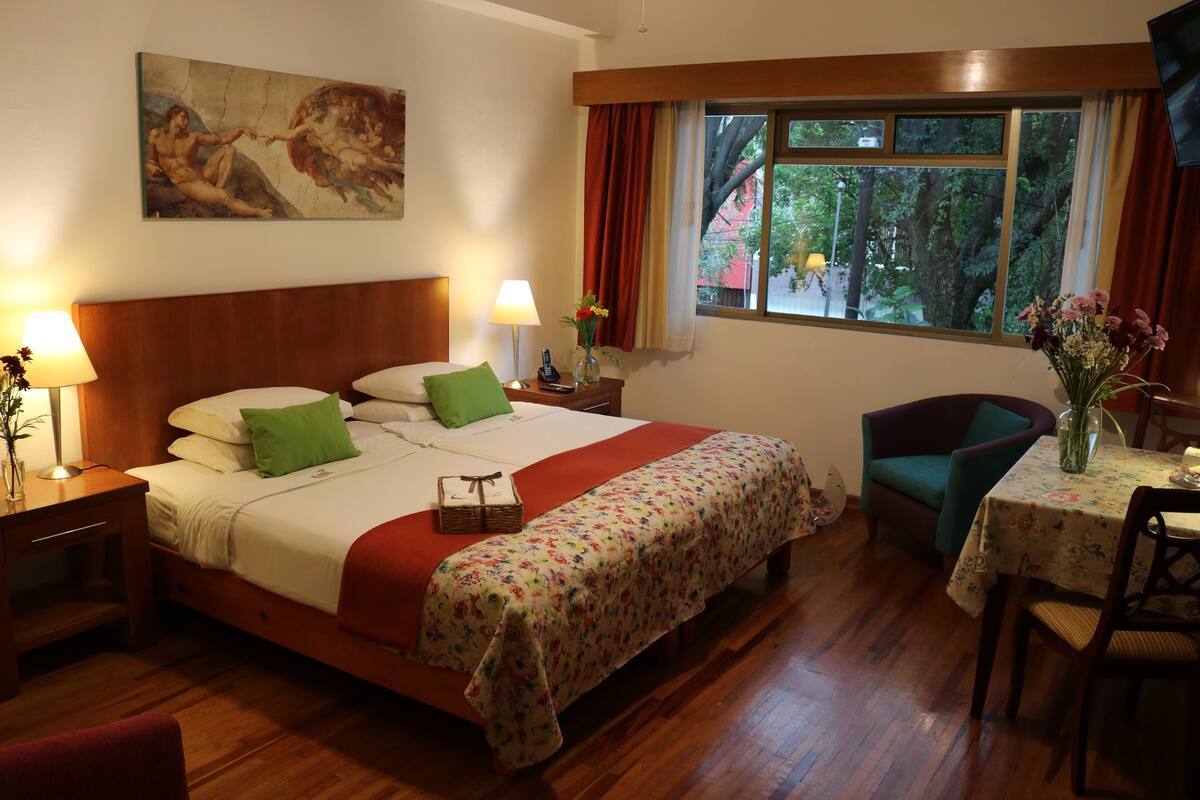 A hotel room with a wooden bed, a desk with a lamp, a chair, a dining table with a vase of flowers, and a window with curtains.