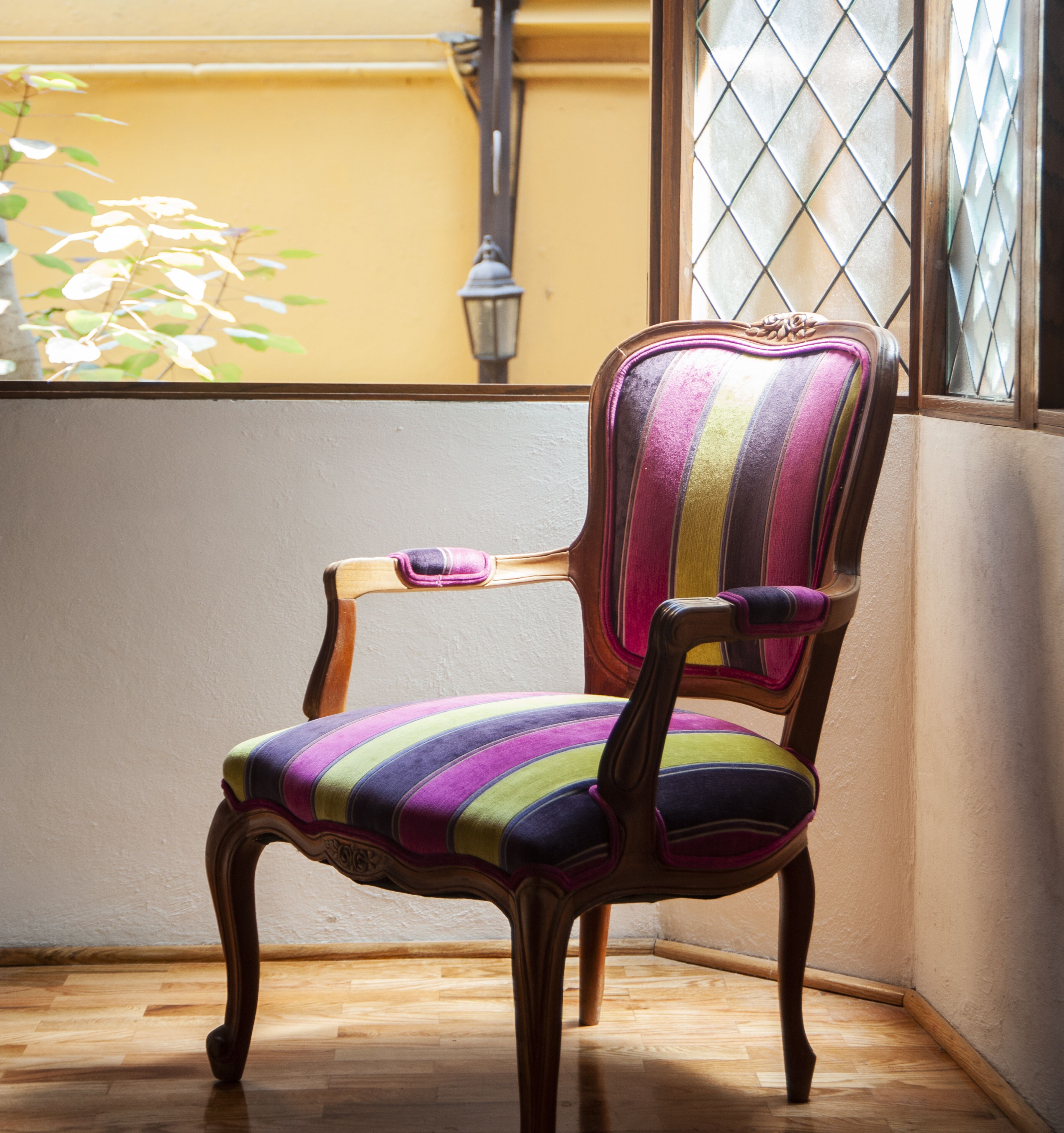 Una silla con un cojín a rayas en un rincón con suelo de madera y una ventana con celosía met álica.