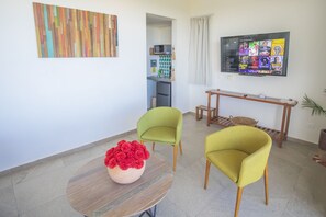 A modern living room with a dining table, two green armchairs, a red flower vase, a kitchen in the background, and a TV mounted on the wall displaying a movie.