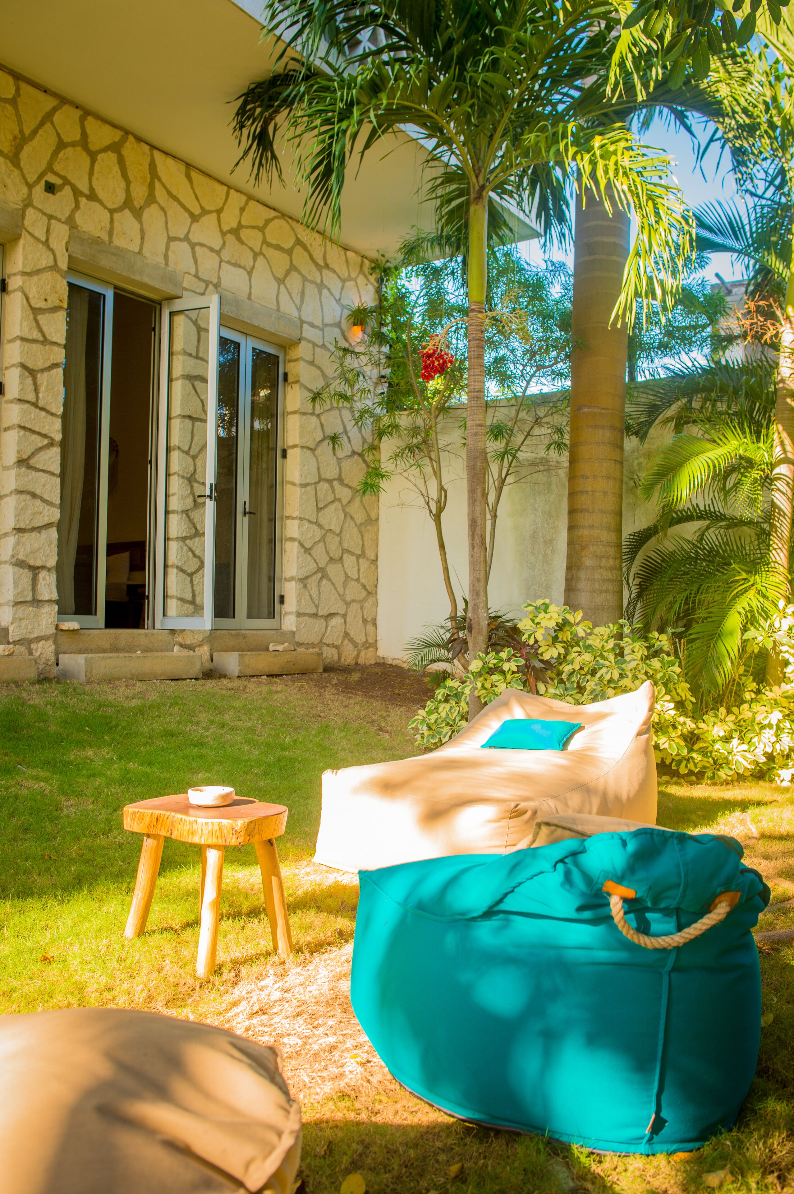 Un edificio de piedra con puerta de vidrio, un taburete de madera y un sillón de saco color turquesa en un área verde con palmeras.