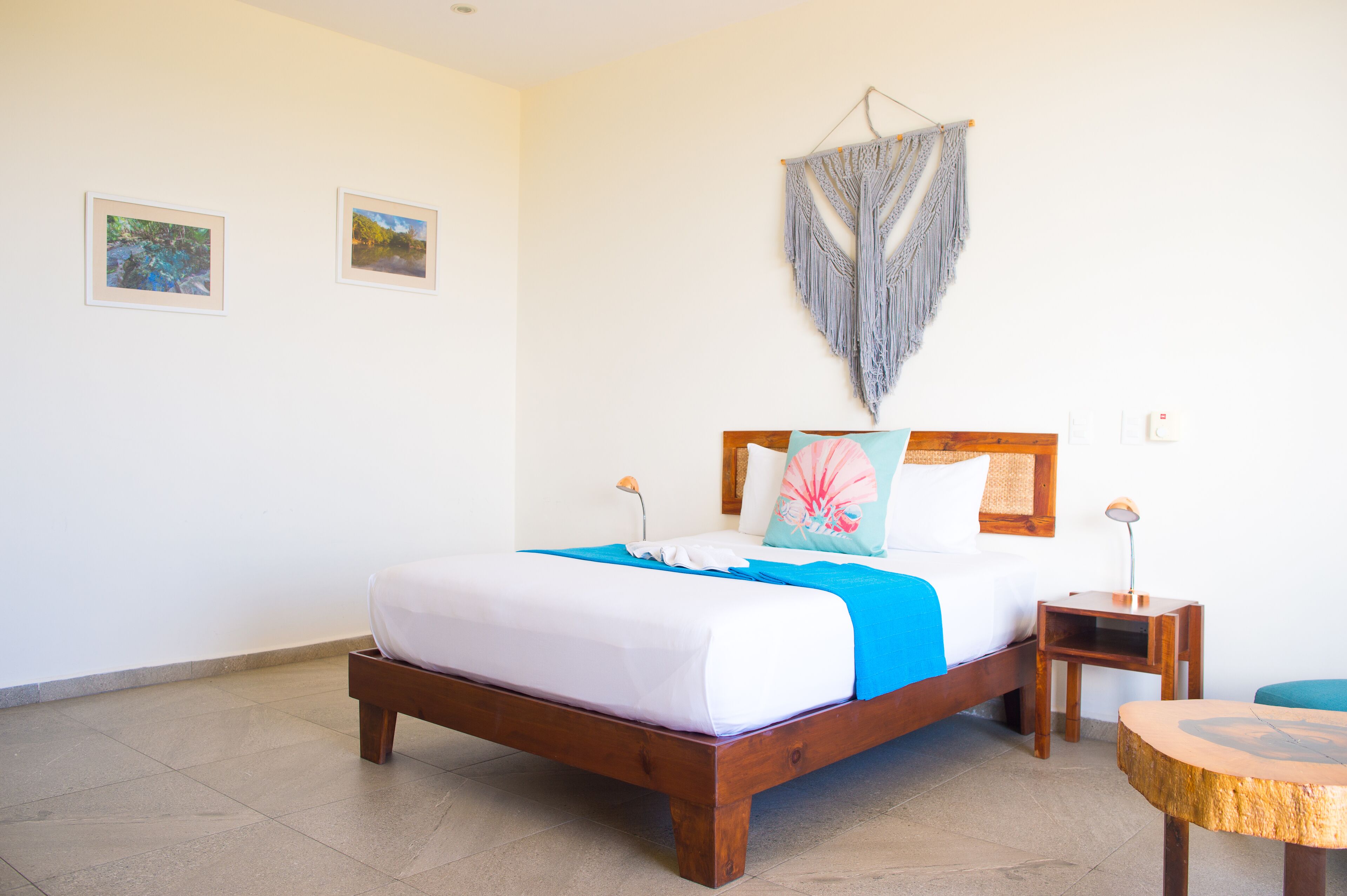 A neatly made bed with a blue and white bedspread, a wooden headboard, and a decorative macrame hanging above. There are two framed pictures on the wall and a small wooden nightstand with a lamp beside the bed.