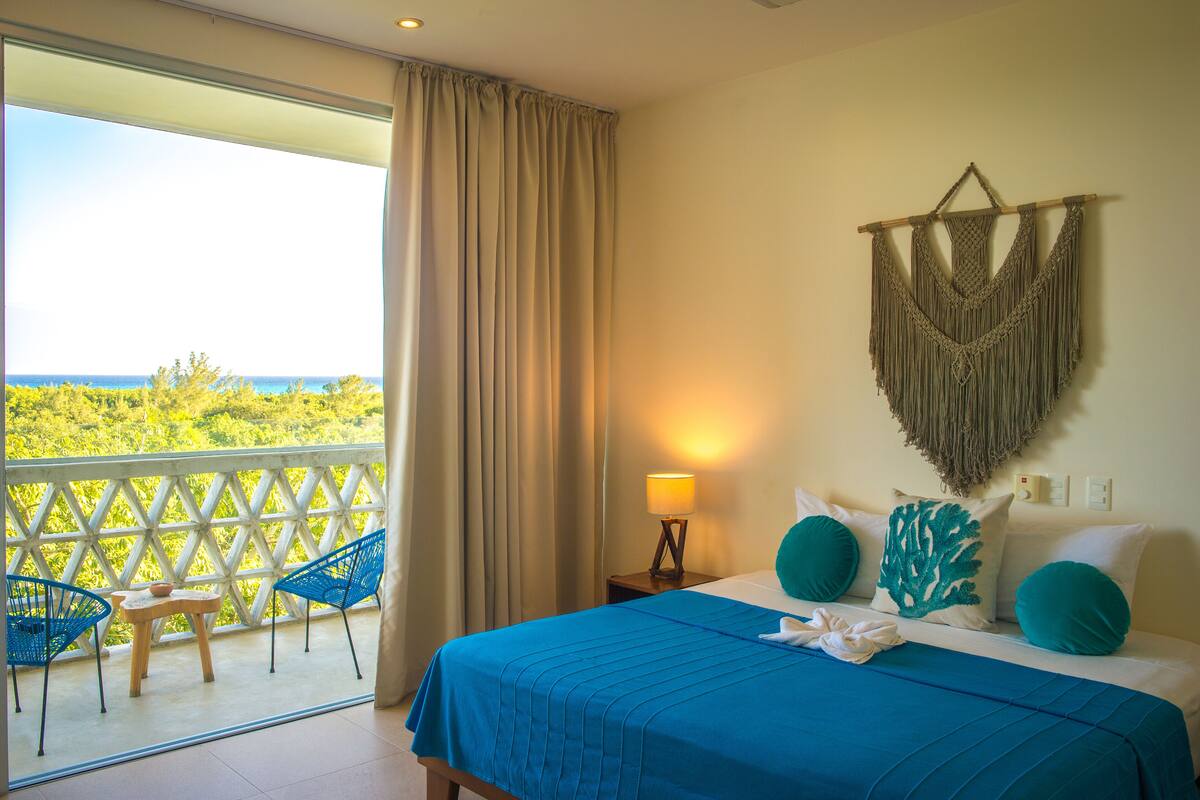 Une chambre d’hôtel avec un balcon, un lit avec une literie bleue et une décoration murale en macramé.